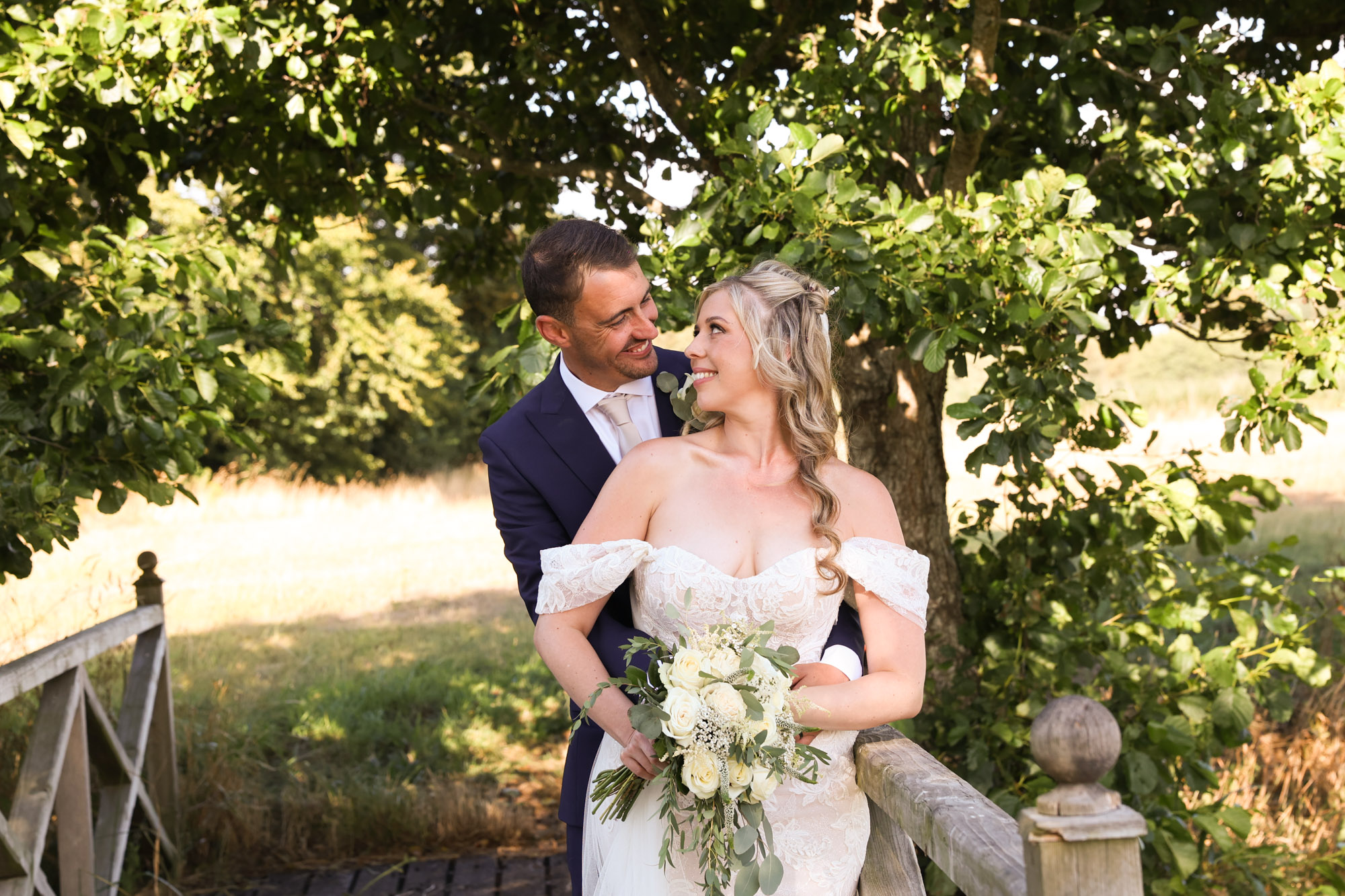 Groom hugging bride from behind, both smiling at each other, standing by a wooden fence with trees in the background.