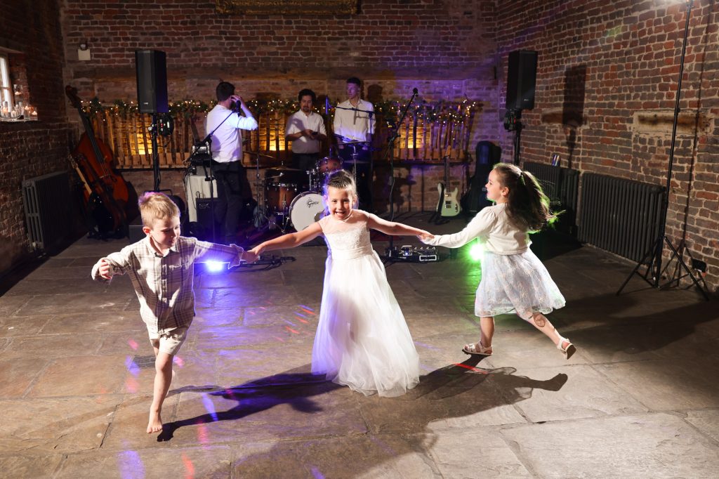 Two young girls and a boy holding hands and dancing on a stone floor in front of a live wedding band.