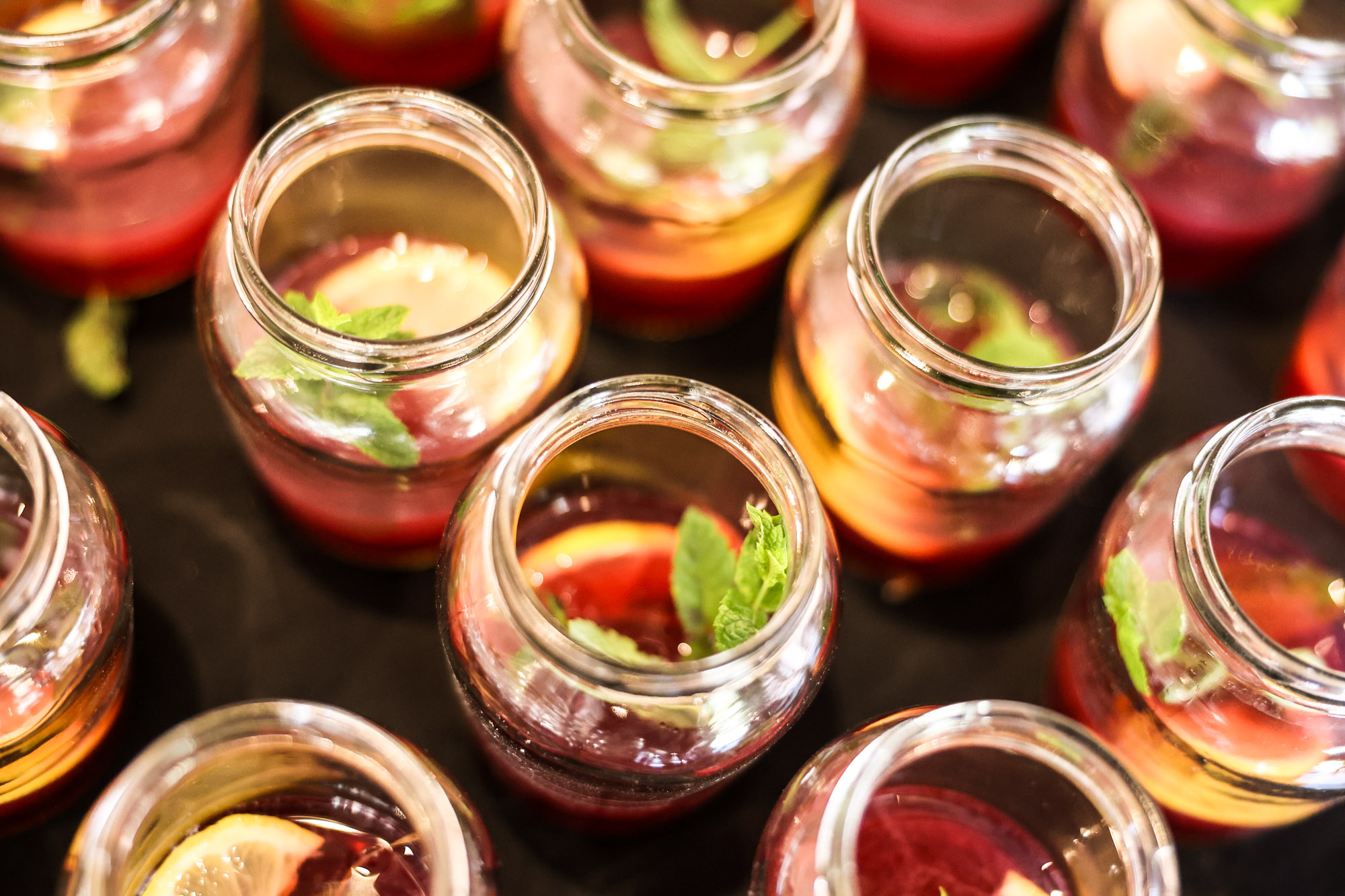Overhead view of multiple small glass jars filled with a red drink, garnished with lemon slices and mint leaves.
