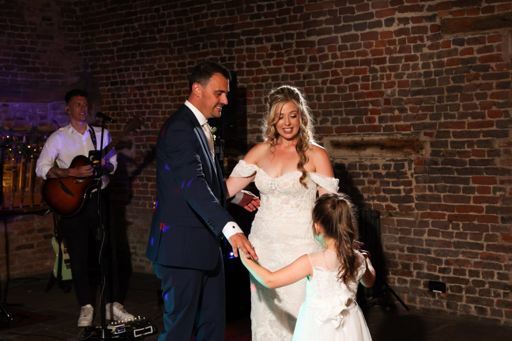 Bride and groom holding hands with a young girl on a dance floor, with a band member playing guitar in the background.