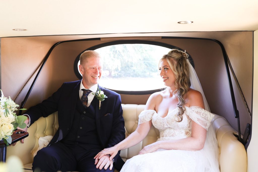 Bride and her father sitting together in the back of a vintage wedding car, smiling at each other.