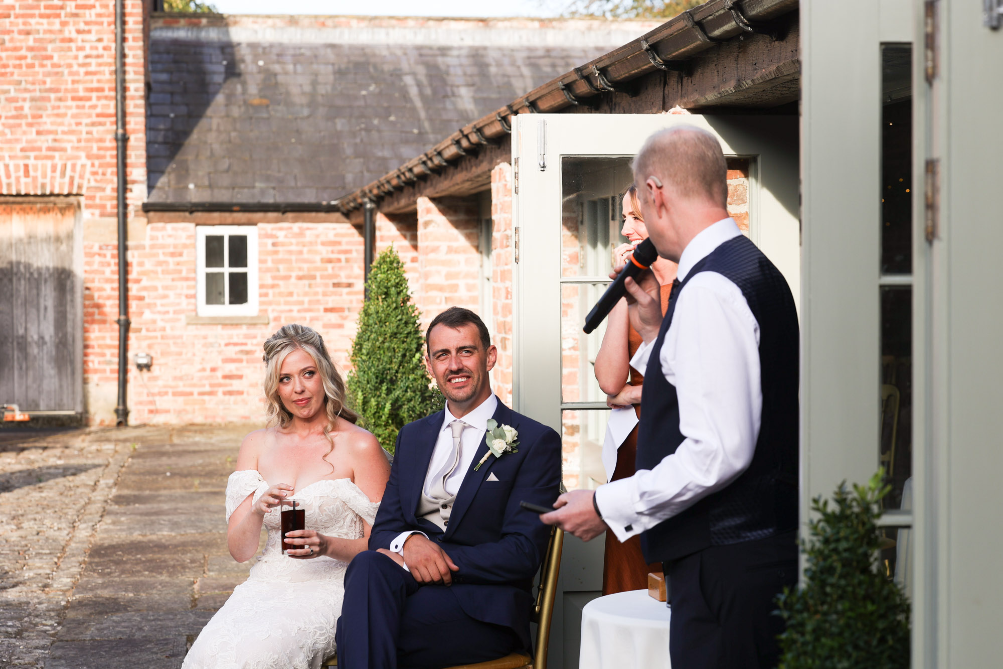 Bride holding a drink and groom seated listening as a bald man in a vest speaks into a microphone outside brick buildings.