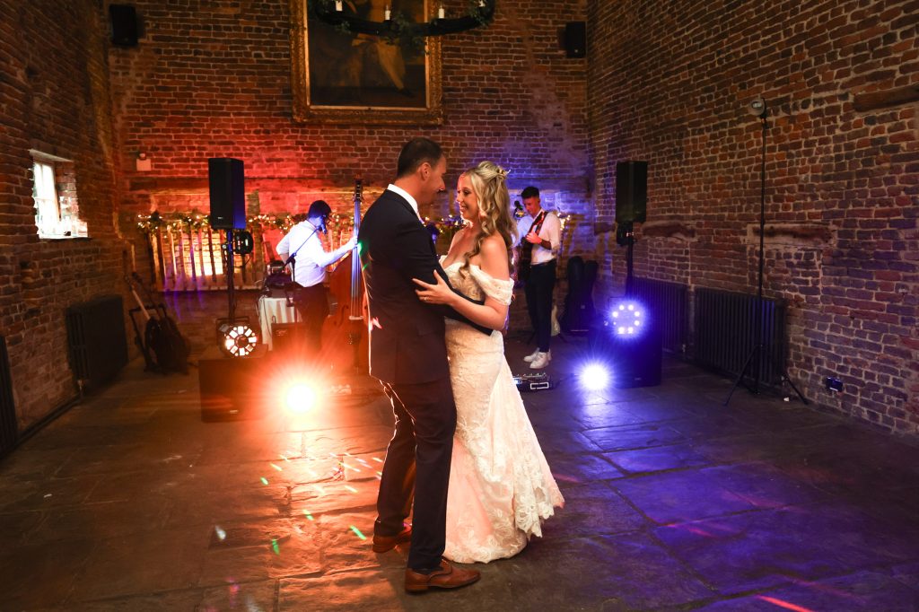 Bride and groom sharing their first dance in a rustic brick hall with colourful stage lighting and a live band behind them.