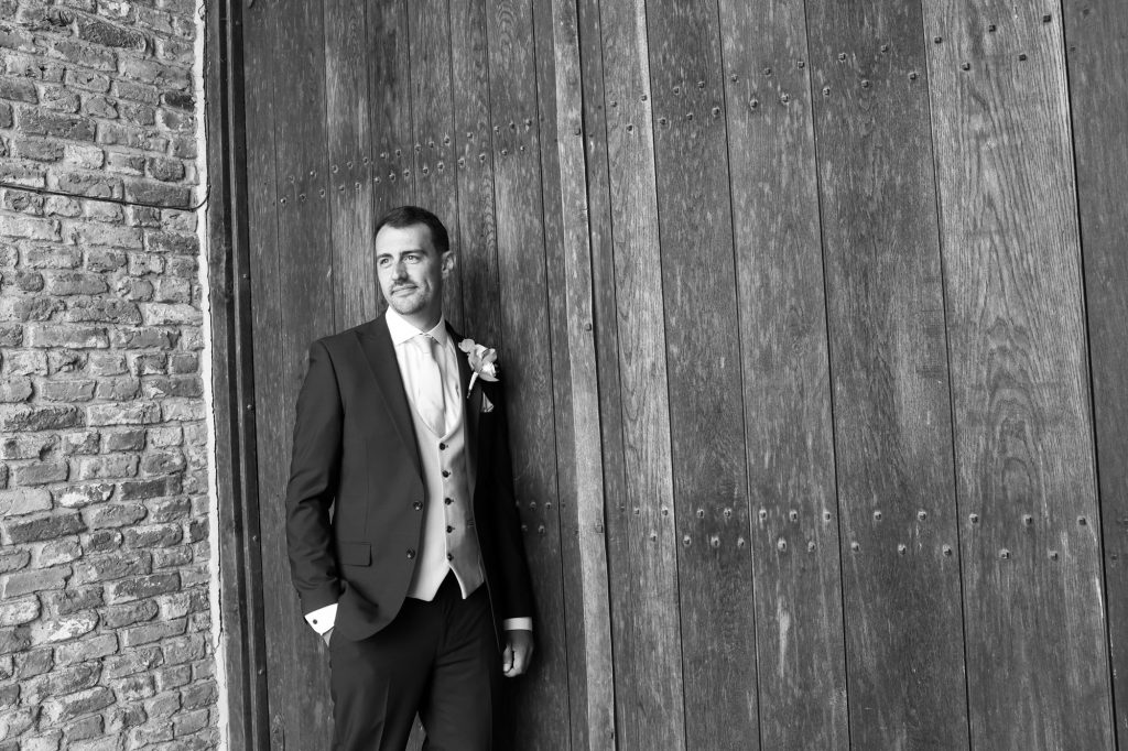Black and white portrait of the groom in a suit standing and looking away against a large wooden barn door.