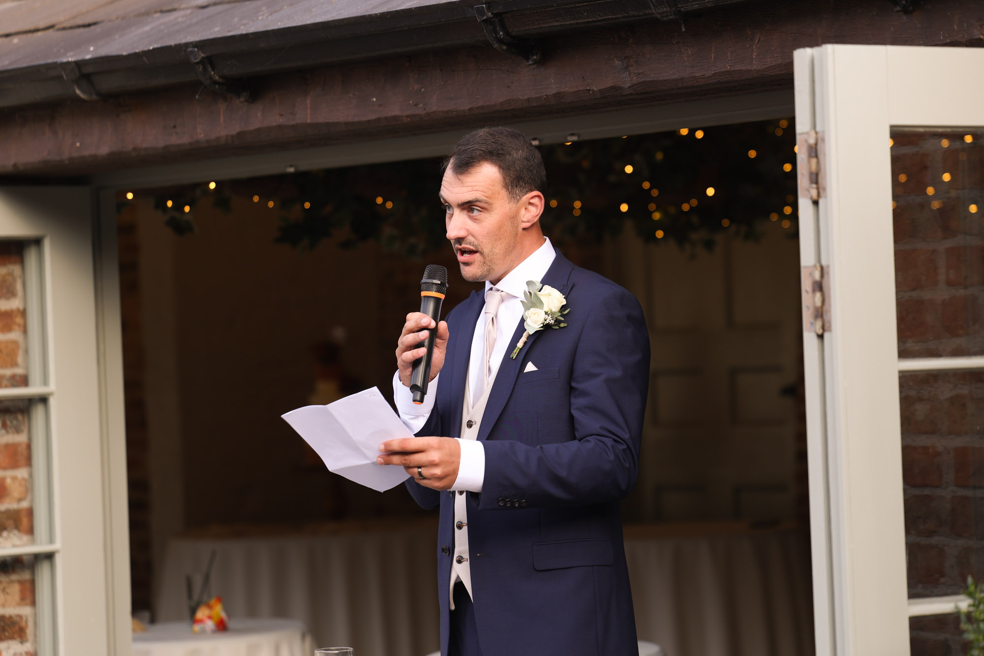 Groom speaking into a microphone and holding speech notes outside in front of open double doors and string lights.