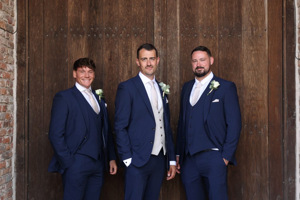 Three men in matching navy blue suits and patterned ties posing together in front of large wooden barn doors.