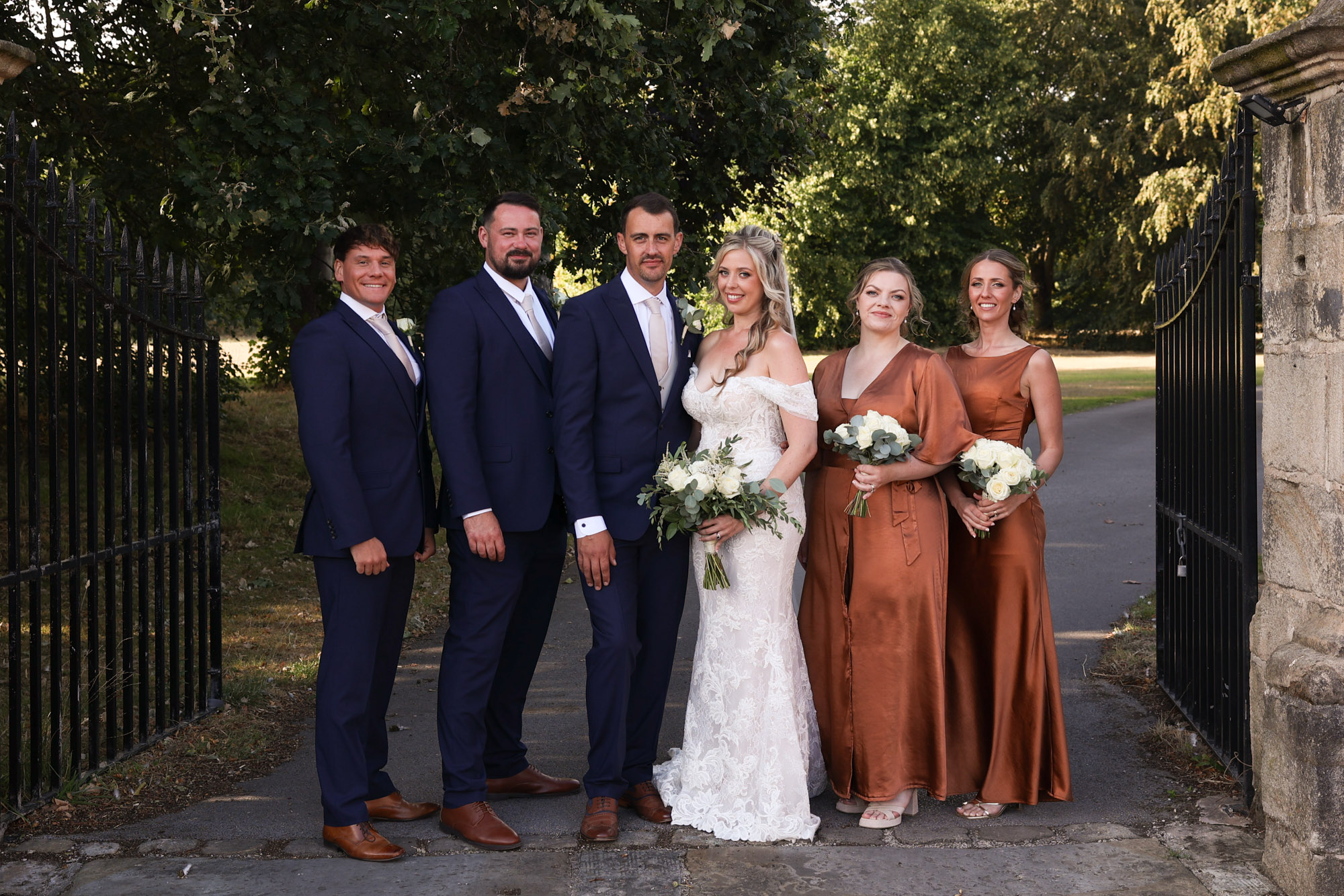 Wedding party of three men in blue suits and three women in rust-orange dresses posing by iron gates and trees.