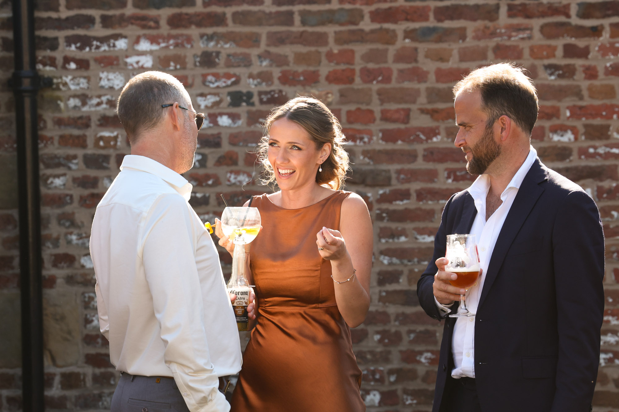 Three wedding guests chatting and smiling together while holding drinks outdoors in front of a brick wall.