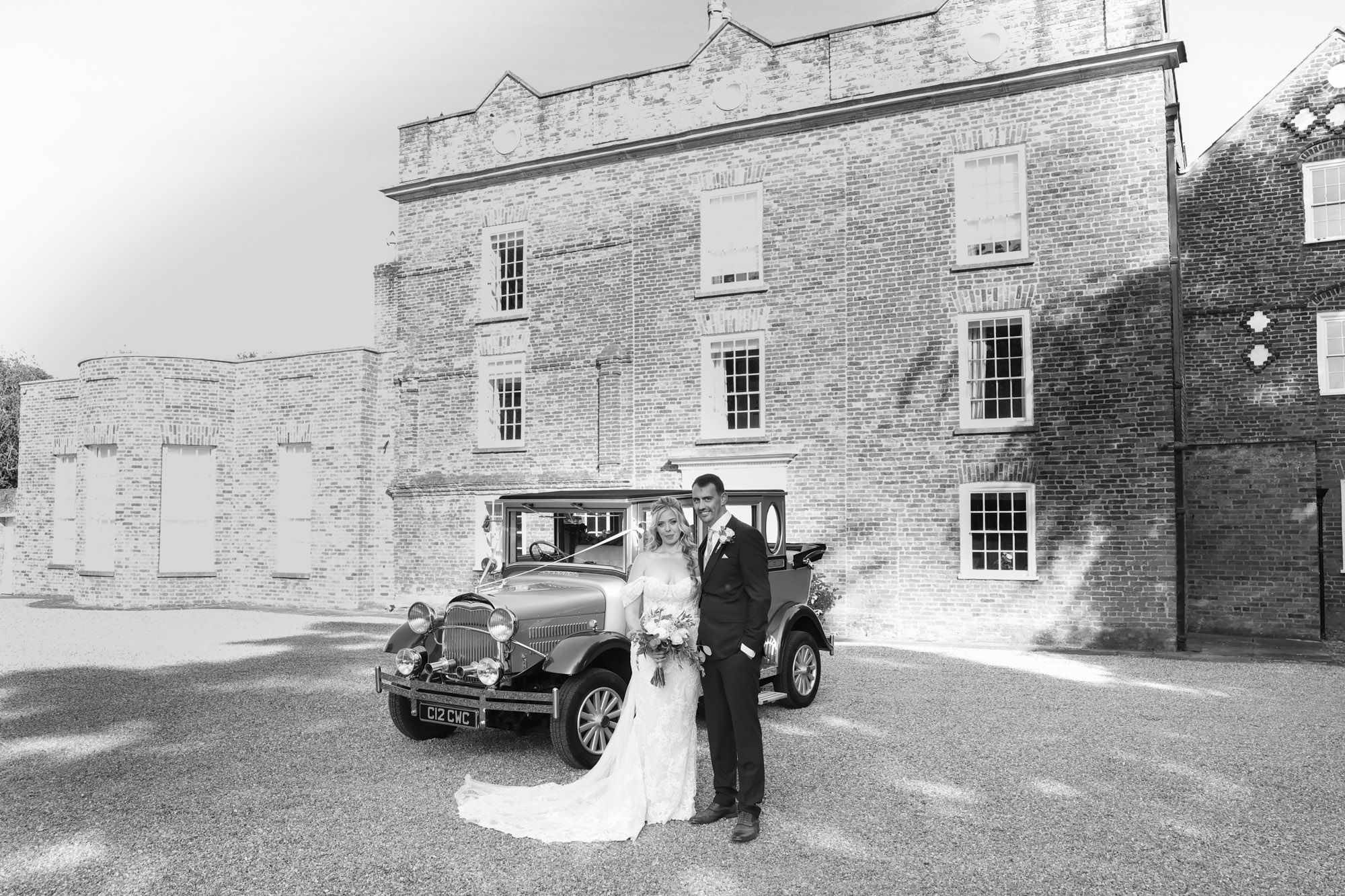 Black and white photo of bride and groom standing beside a vintage car in front of a historic brick manor house.