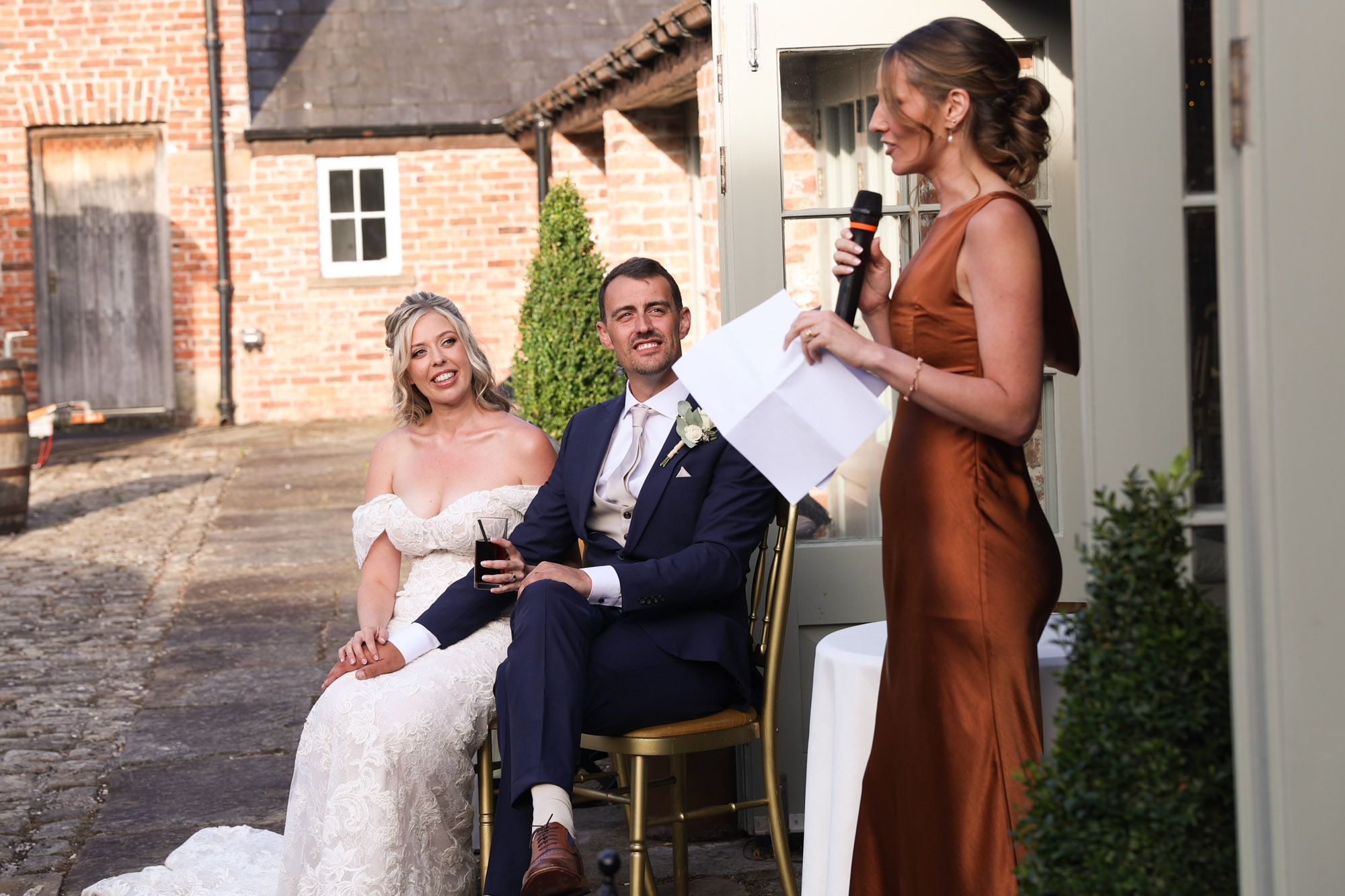 Maid of honour in a rust-orange dress giving a speech with a microphone, with the bride and groom seated behind her.