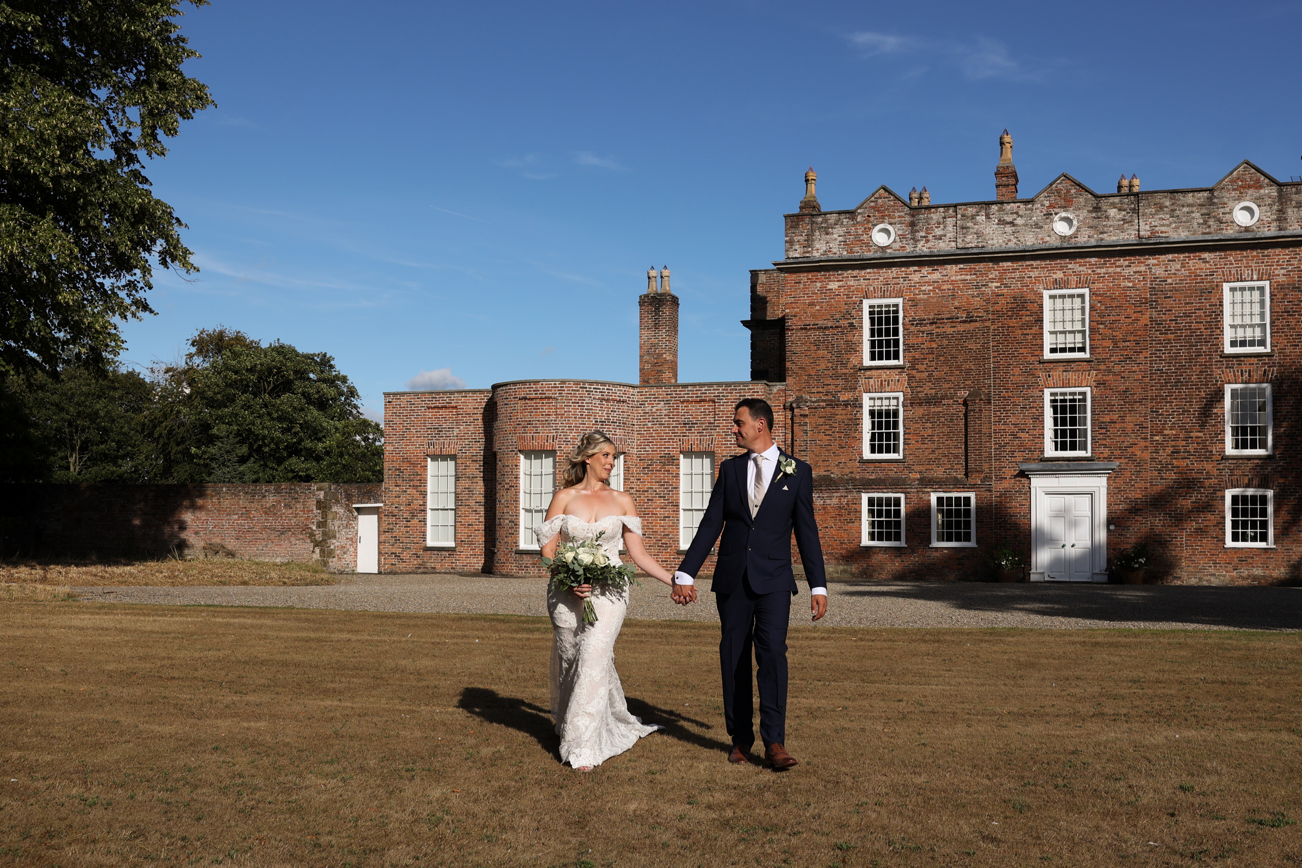 Bride and groom holding hands and walking together across a sunny lawn in front of a grand brick manor house.