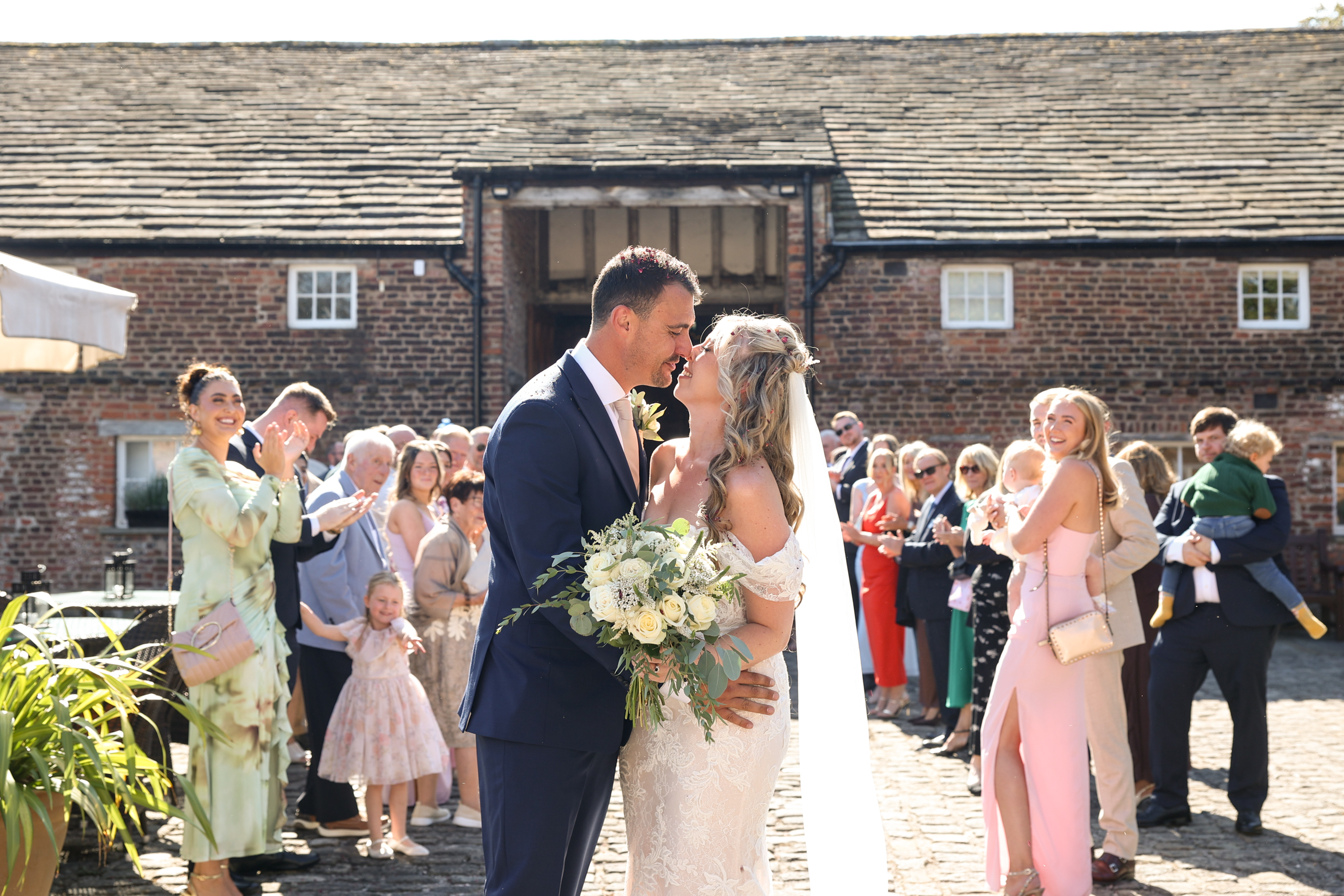 Newlywed couple leaning in for a kiss outside a rustic brick building while cheering guests throw confetti.