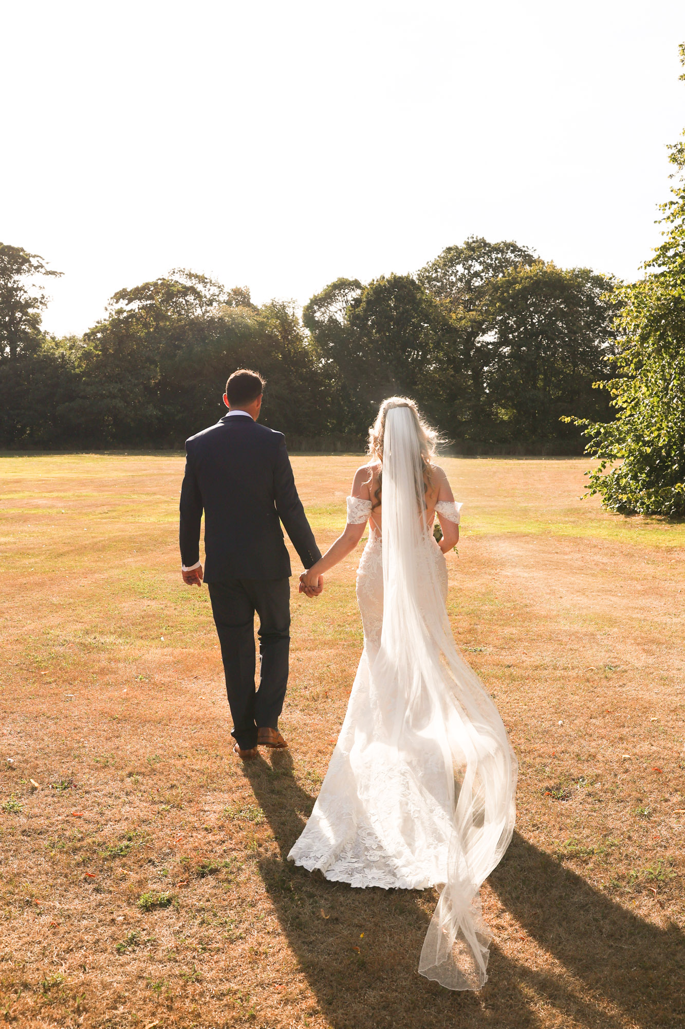 Bride and groom holding hands and walking together across a sunny grass field with trees in the background.