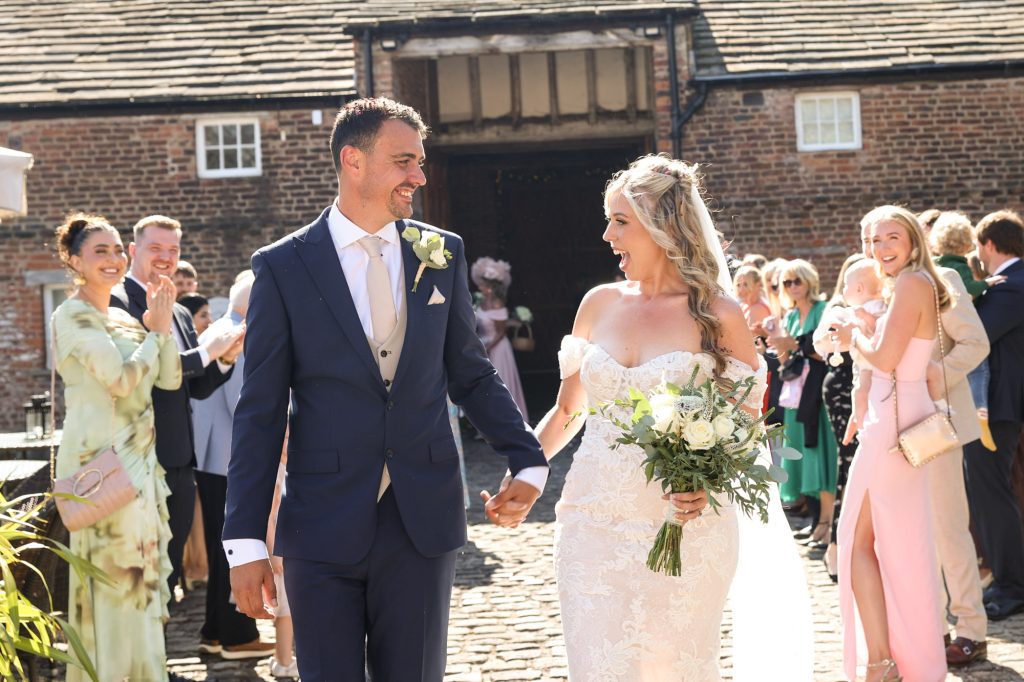 Newlywed couple smiling and holding hands while walking outside a brick barn amidst cheering guests.