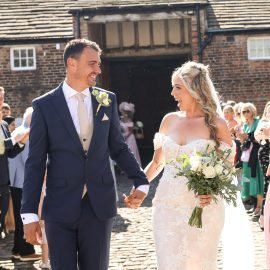 Newlywed couple smiling and holding hands while walking outside a brick barn amidst cheering guests.