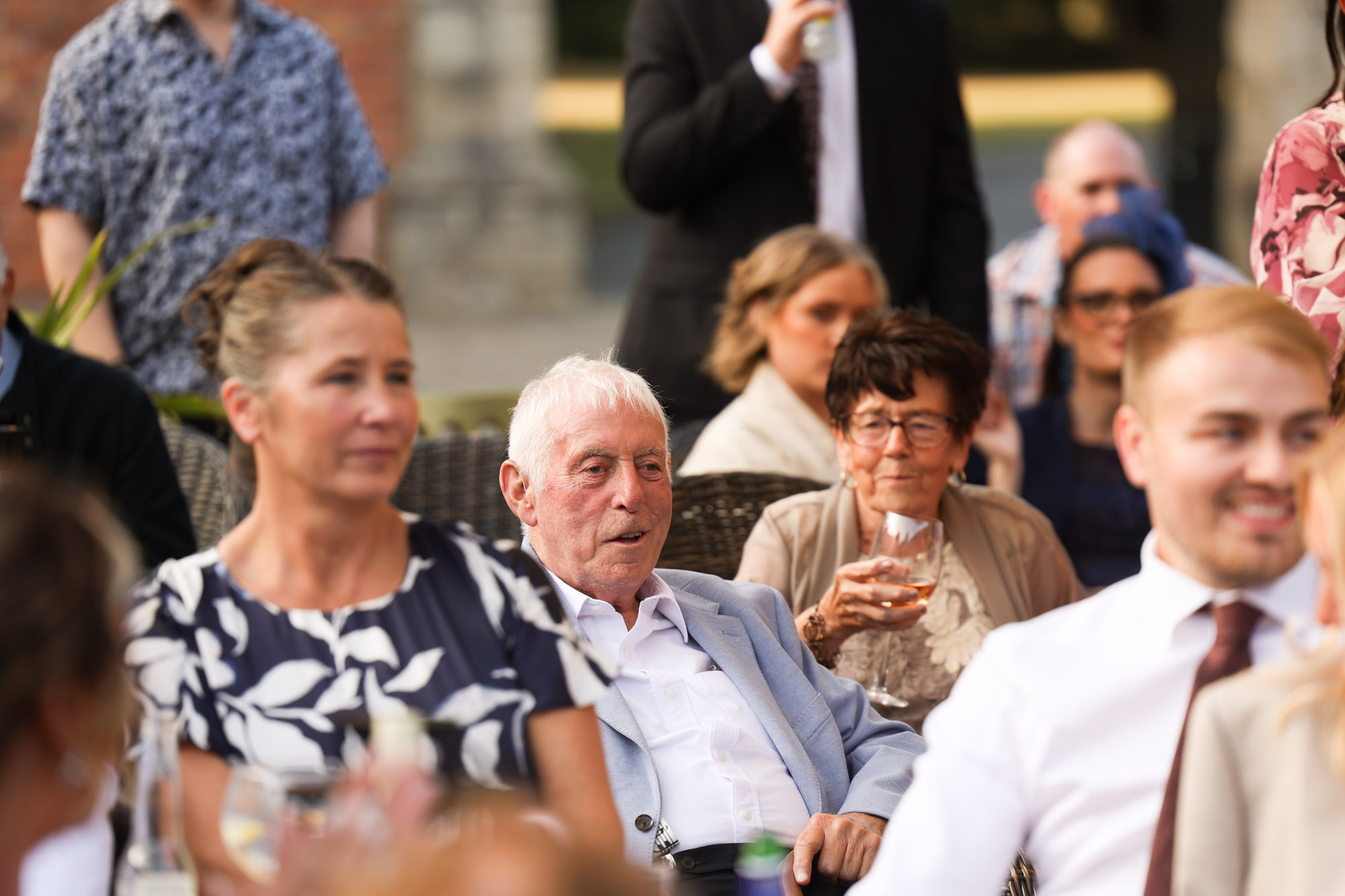 Older man with white hair seated and looking forward among wedding guests during speeches outdoors.