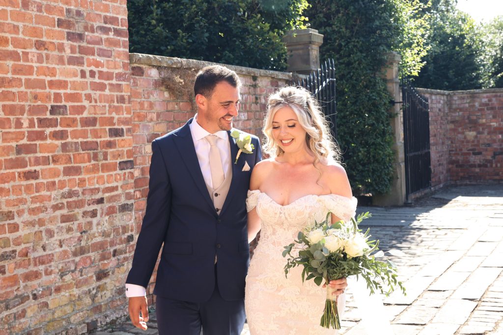 Groom and bride smiling at each other while walking together along a sunny path beside a brick wall.