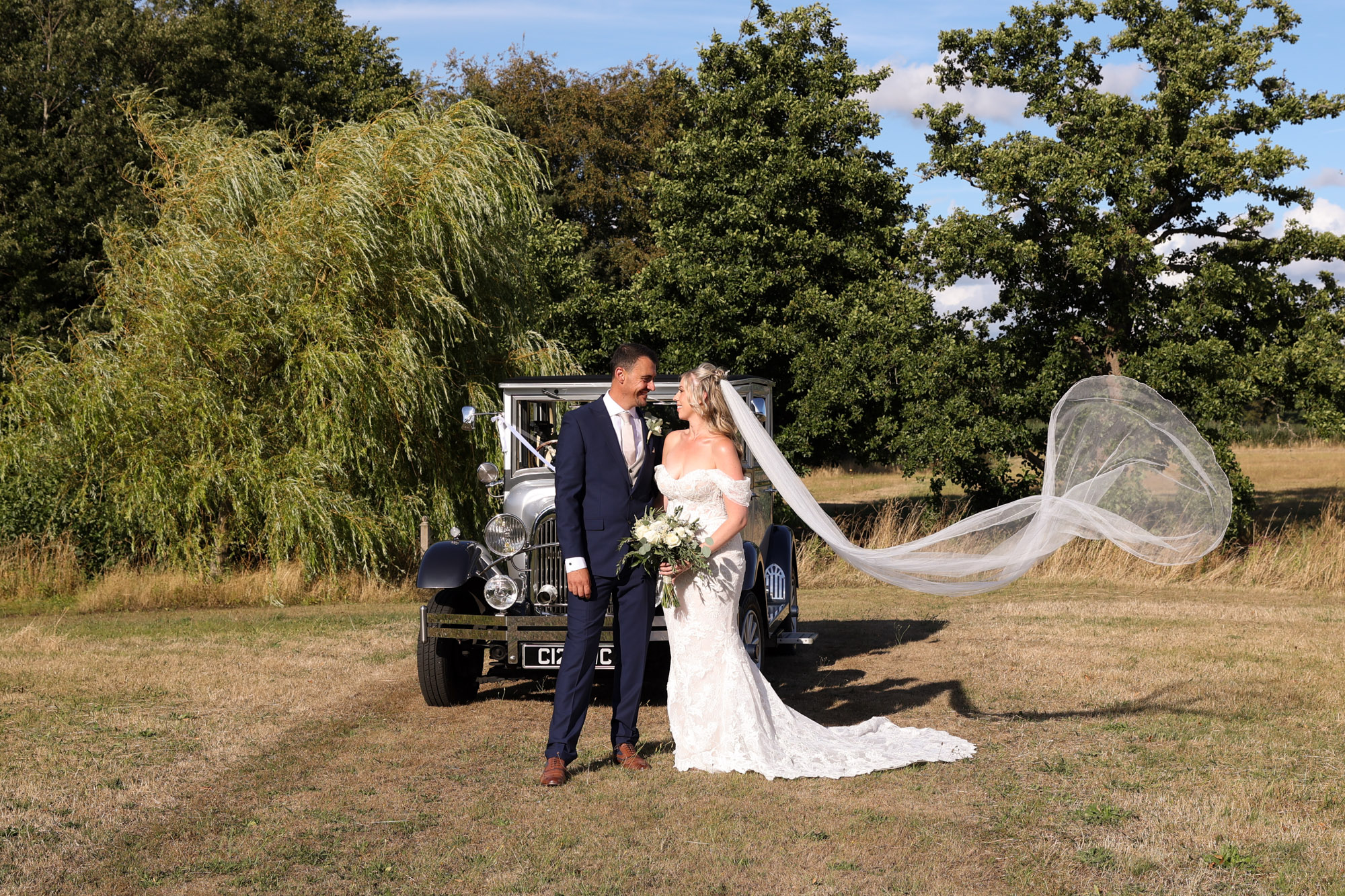 Bride and groom standing by a vintage car on a grassy field with a large veil billowing in the wind.