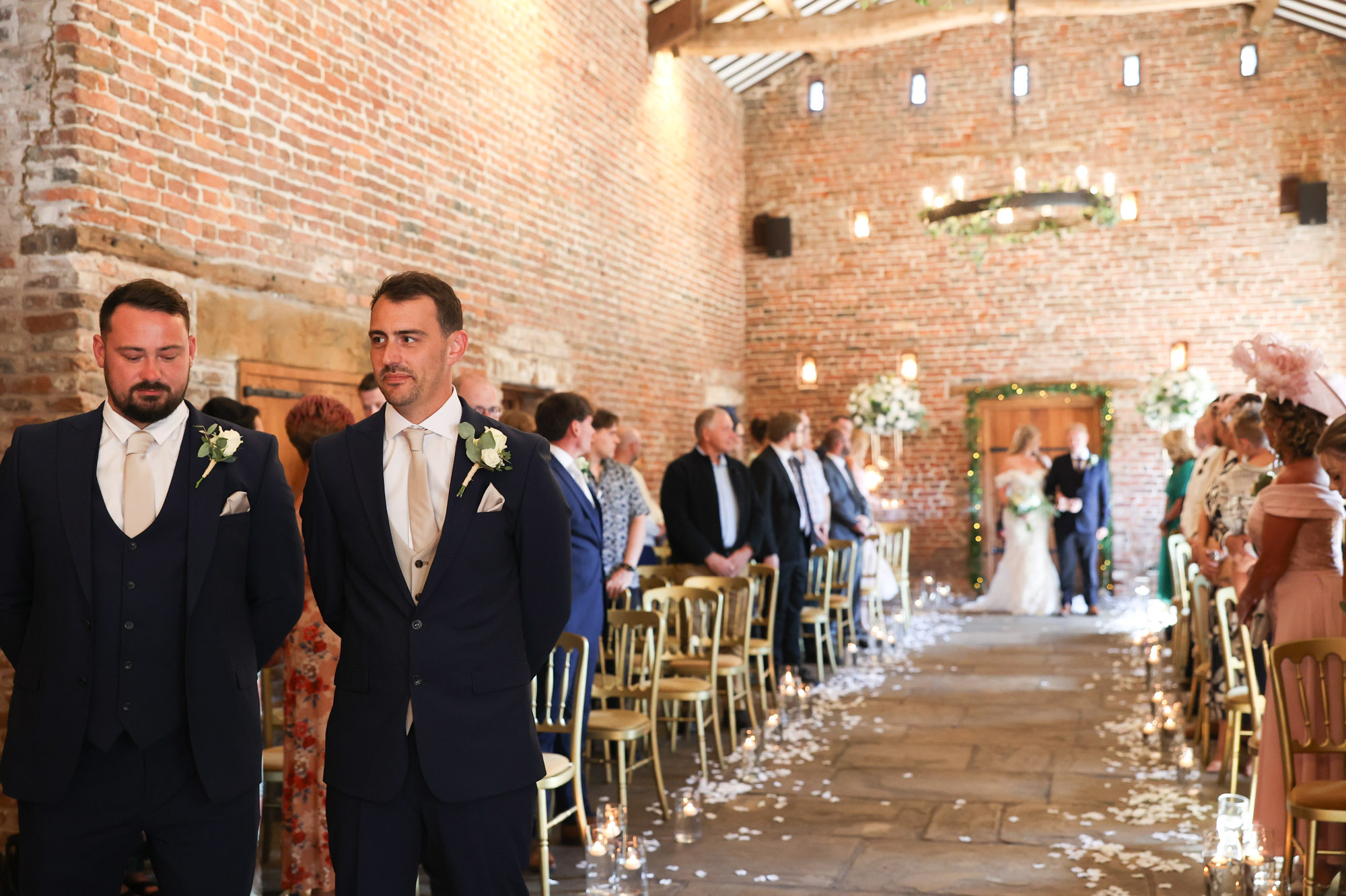 Groom and groomsmen standing at the front of a rustic brick barn waiting as the bride walks down the aisle.