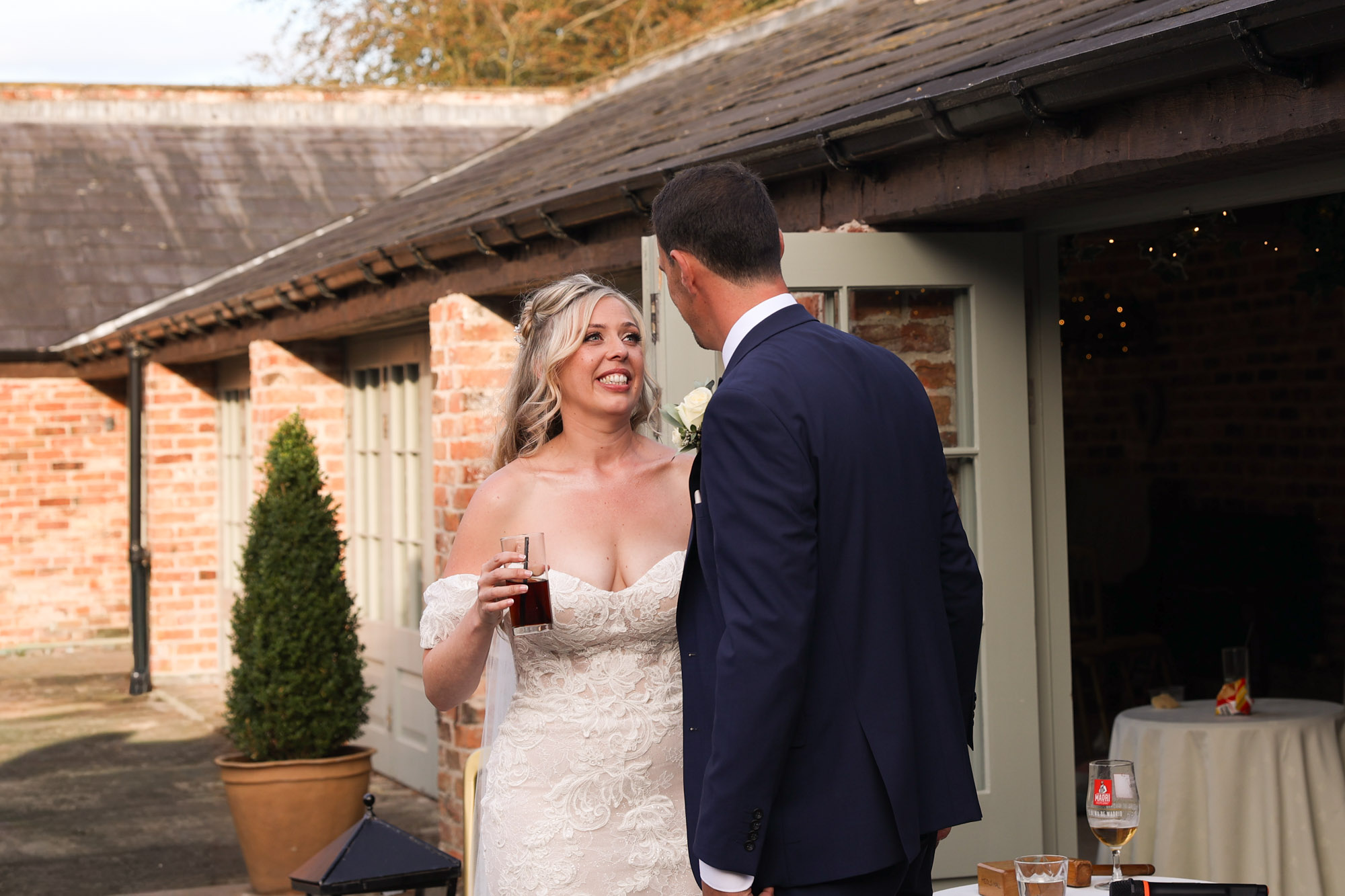 Bride in a lace wedding gown holding a dark drink, smiling at the groom outdoors.