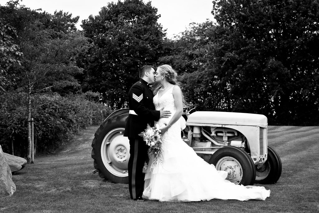 Black and white photo of a bride and groom kissing next to a vintage tractor outdoors.