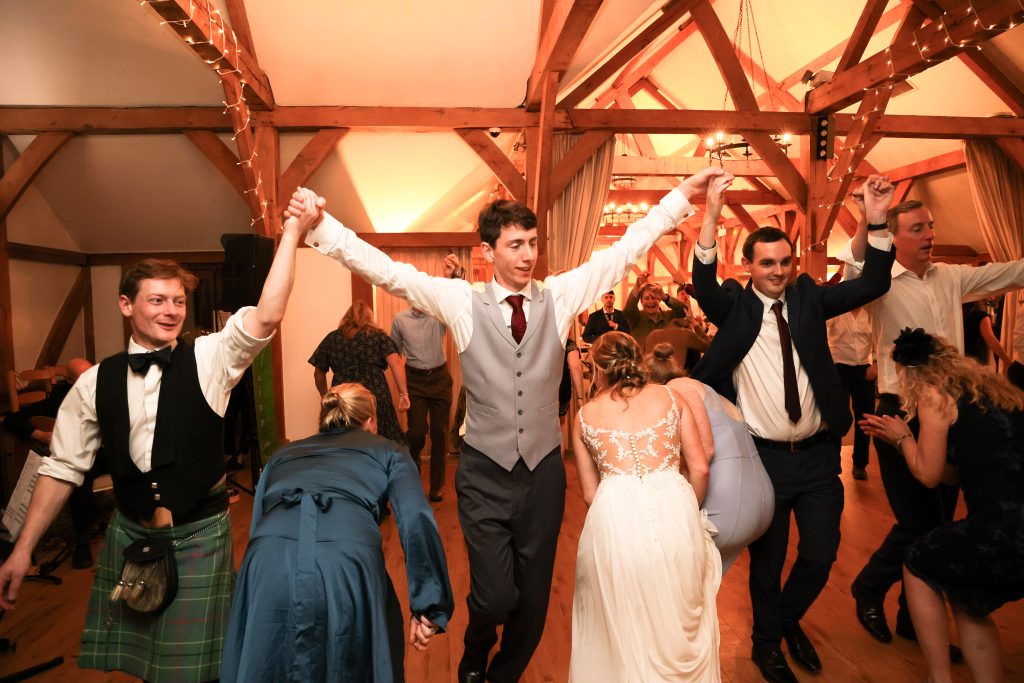 Wedding guests dancing and holding hands during a lively ceilidh in the barn.