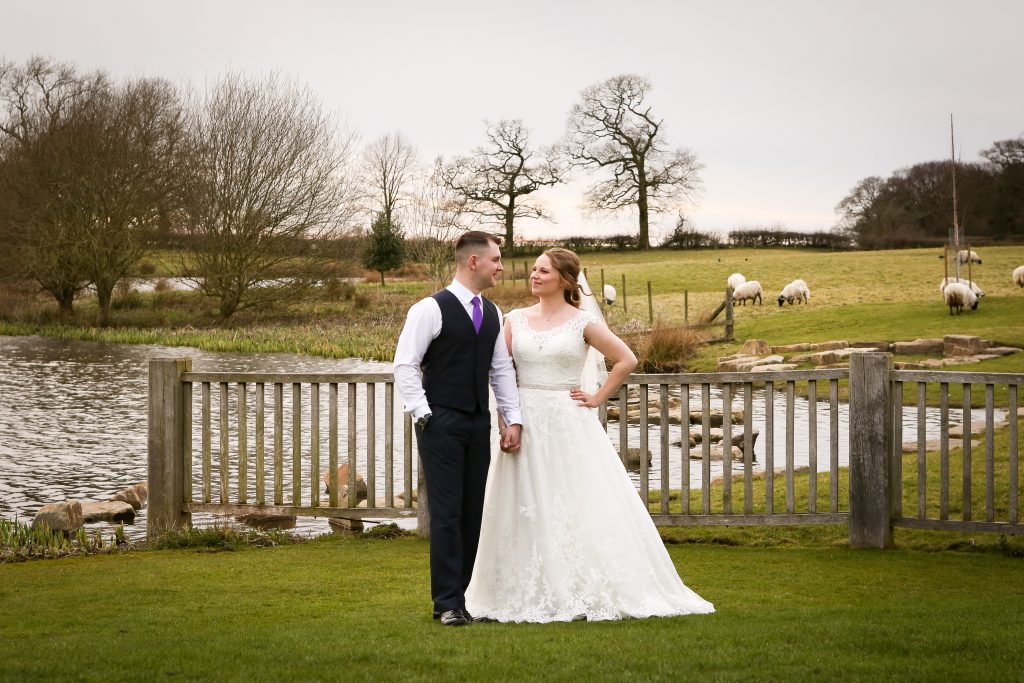 Bride and groom standing by a wooden fence with sheep grazing in the countryside.