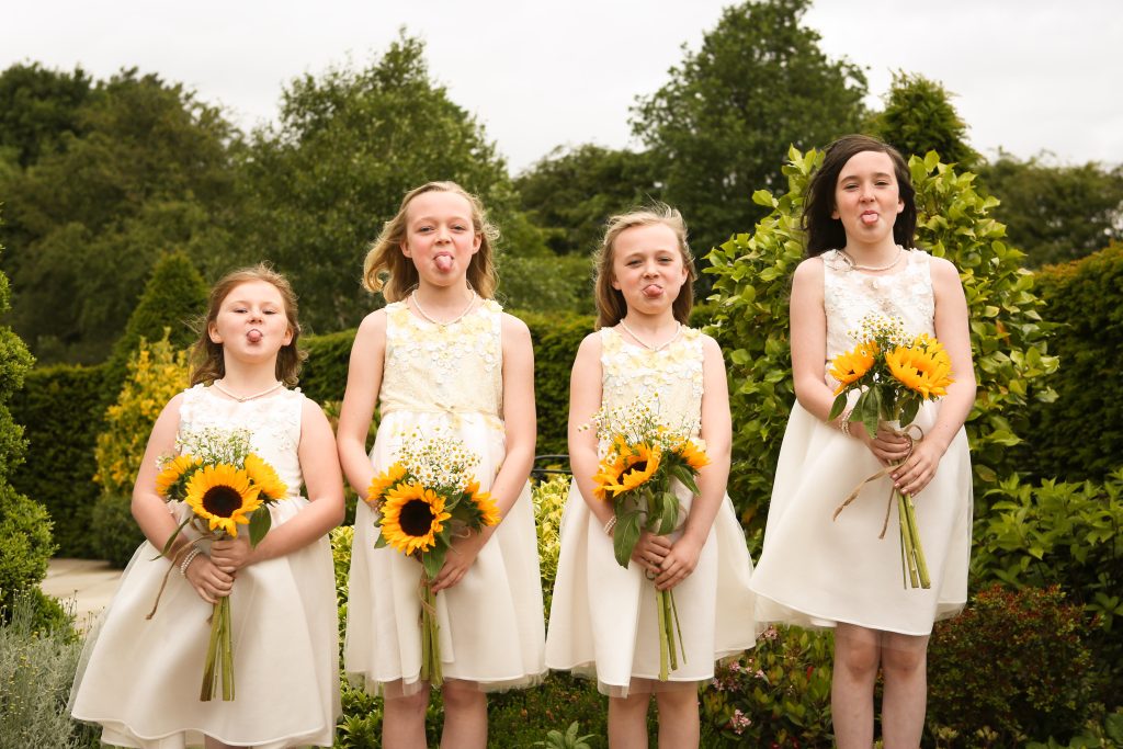 Four flower girls in white dresses holding sunflower bouquets and sticking their tongues out in a garden.