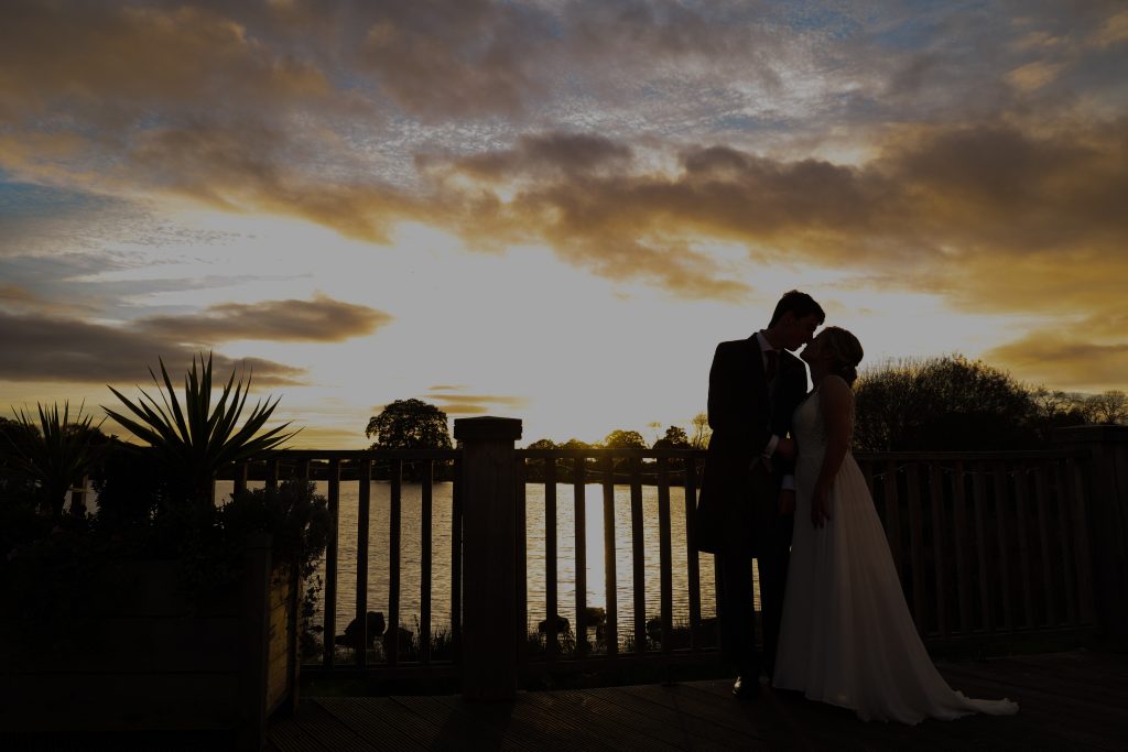 Silhouette of a bride and groom kissing on a wooden balcony against a dramatic sunset.