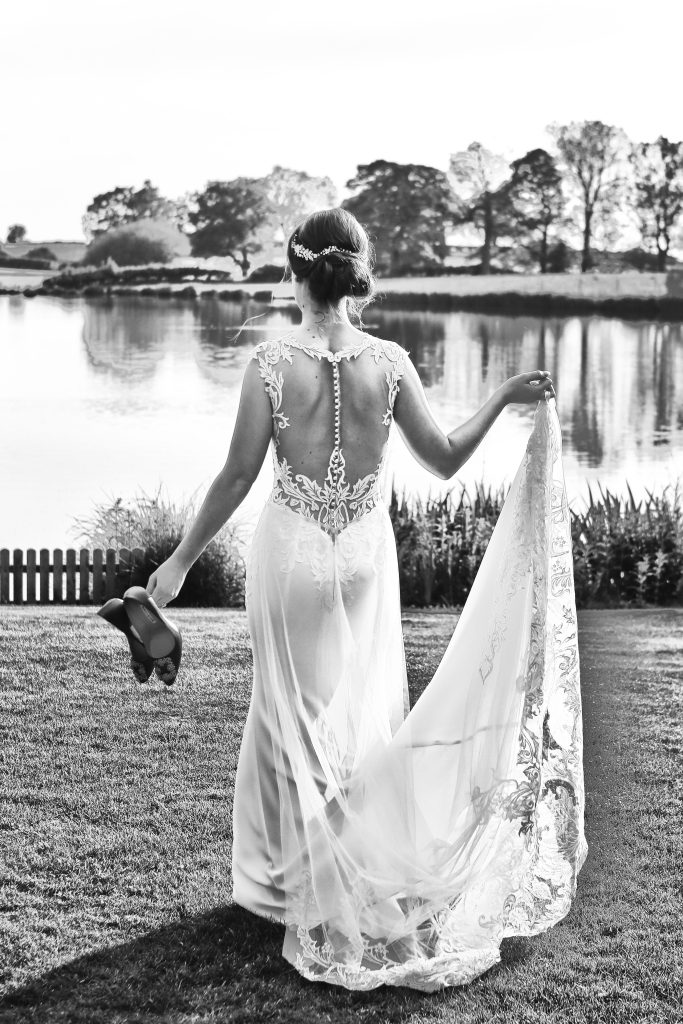 Black and white back view of a bride walking by a lake holding her shoes and train.