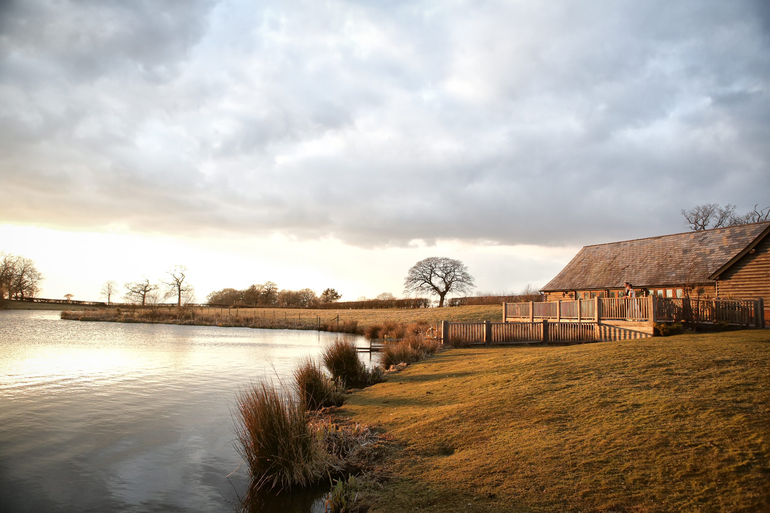 Wide view of Sandhole Oak Barn and its scenic lake under a cloudy sunset sky.