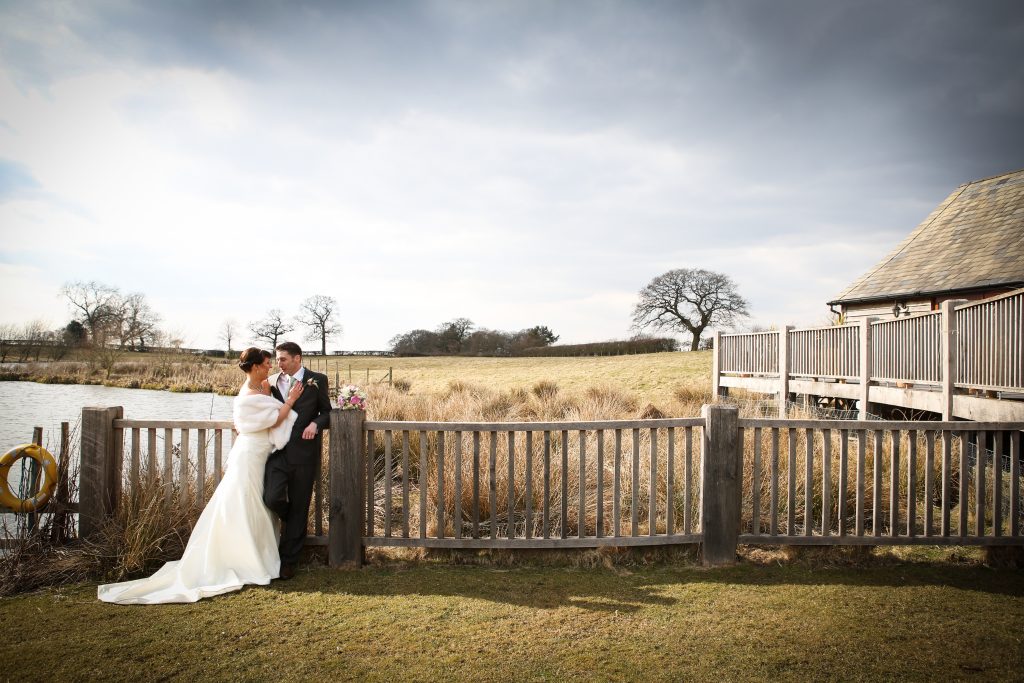 Bride and groom posing by a wooden fence and lake outside Sandhole Oak Barn.