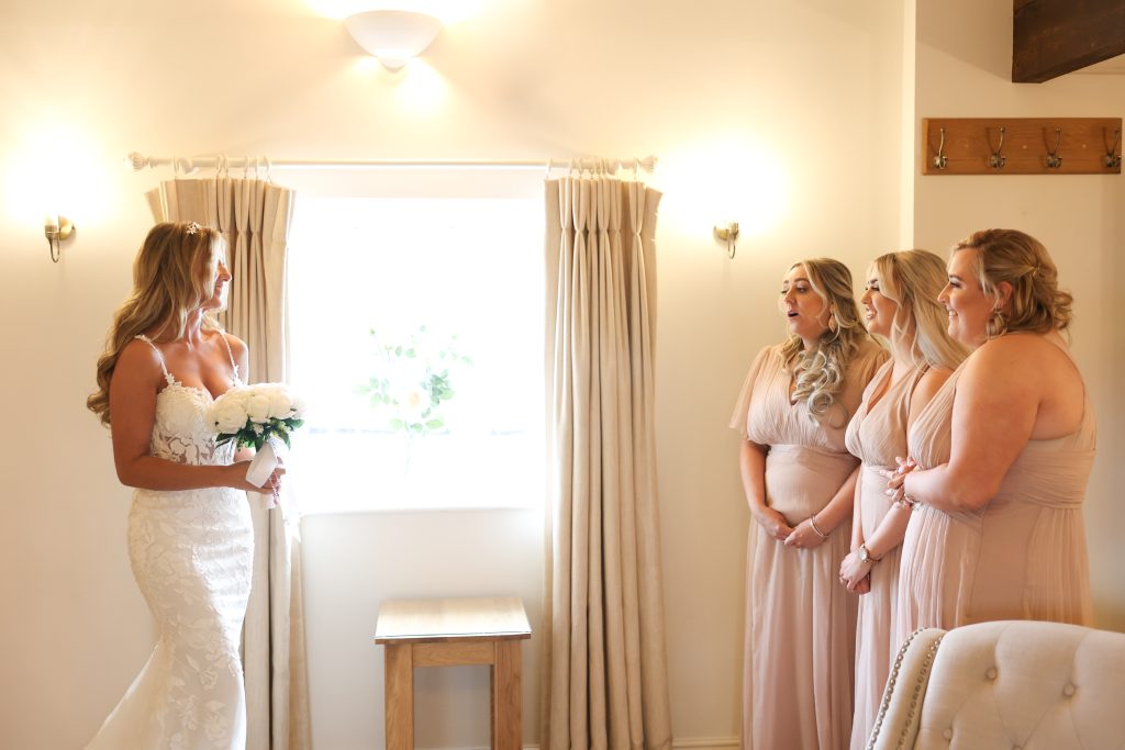 Bride holding white flowers while looking at her bridesmaids in a bright room.