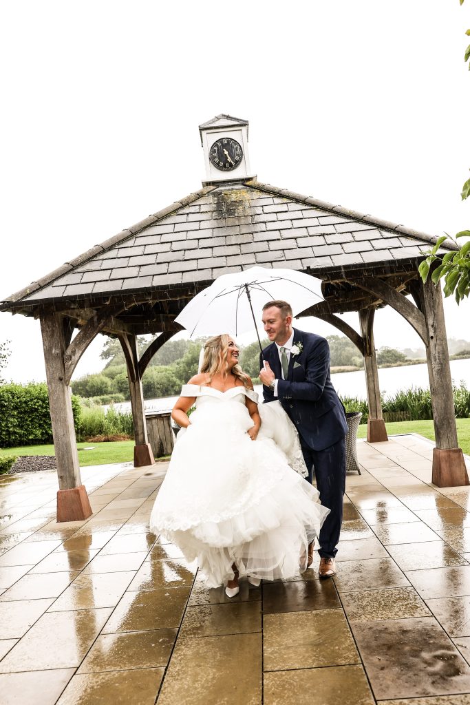 Bride and groom laughing under a white umbrella near the lakeside gazebo in the rain.