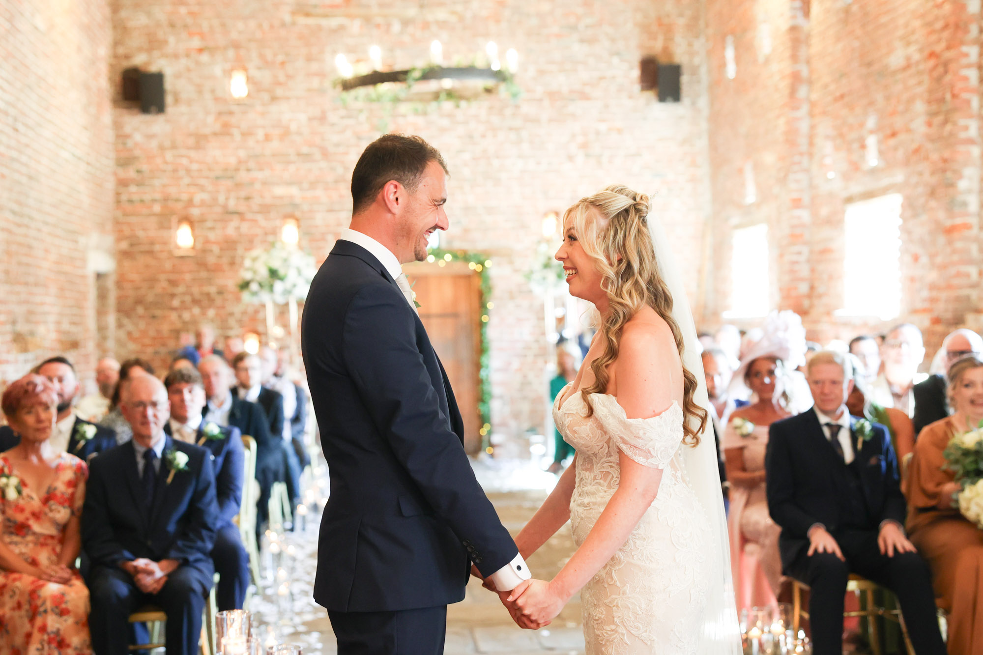 Bride and groom smiling at each other and holding hands during their wedding ceremony inside a brick barn.