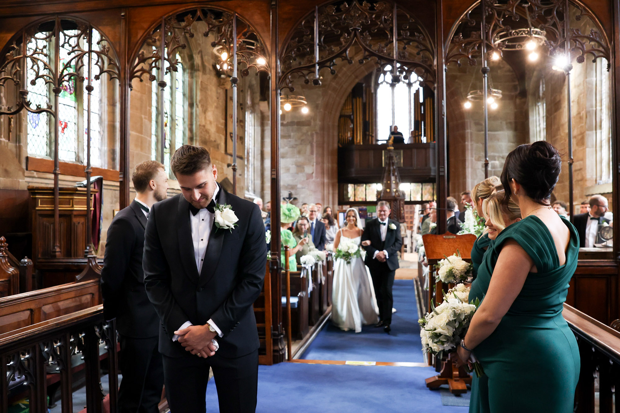 Groom in tuxedo waits looking down as bride walks down church aisle with father.