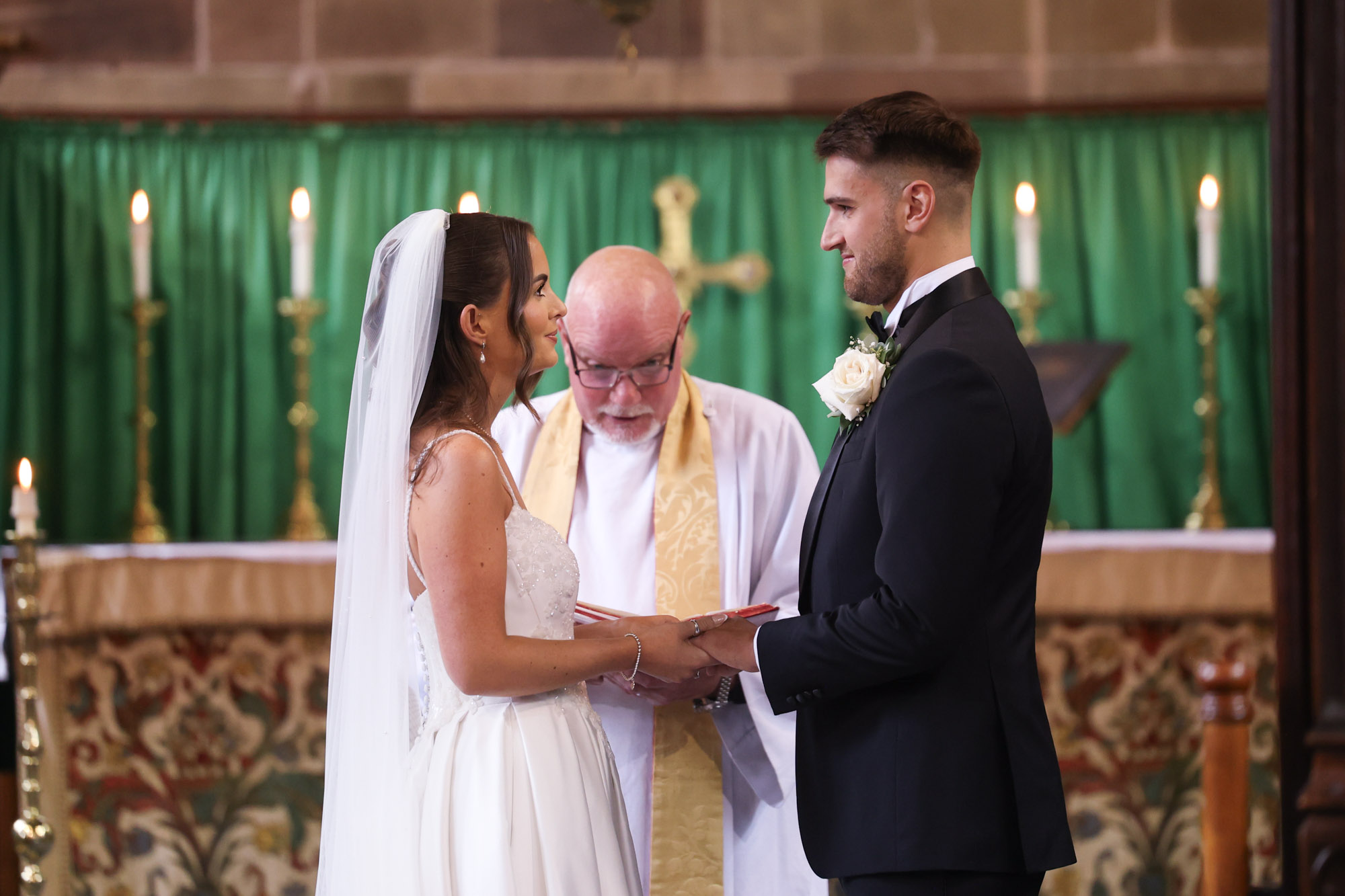 Bride and groom hold hands facing priest during church wedding ceremony.