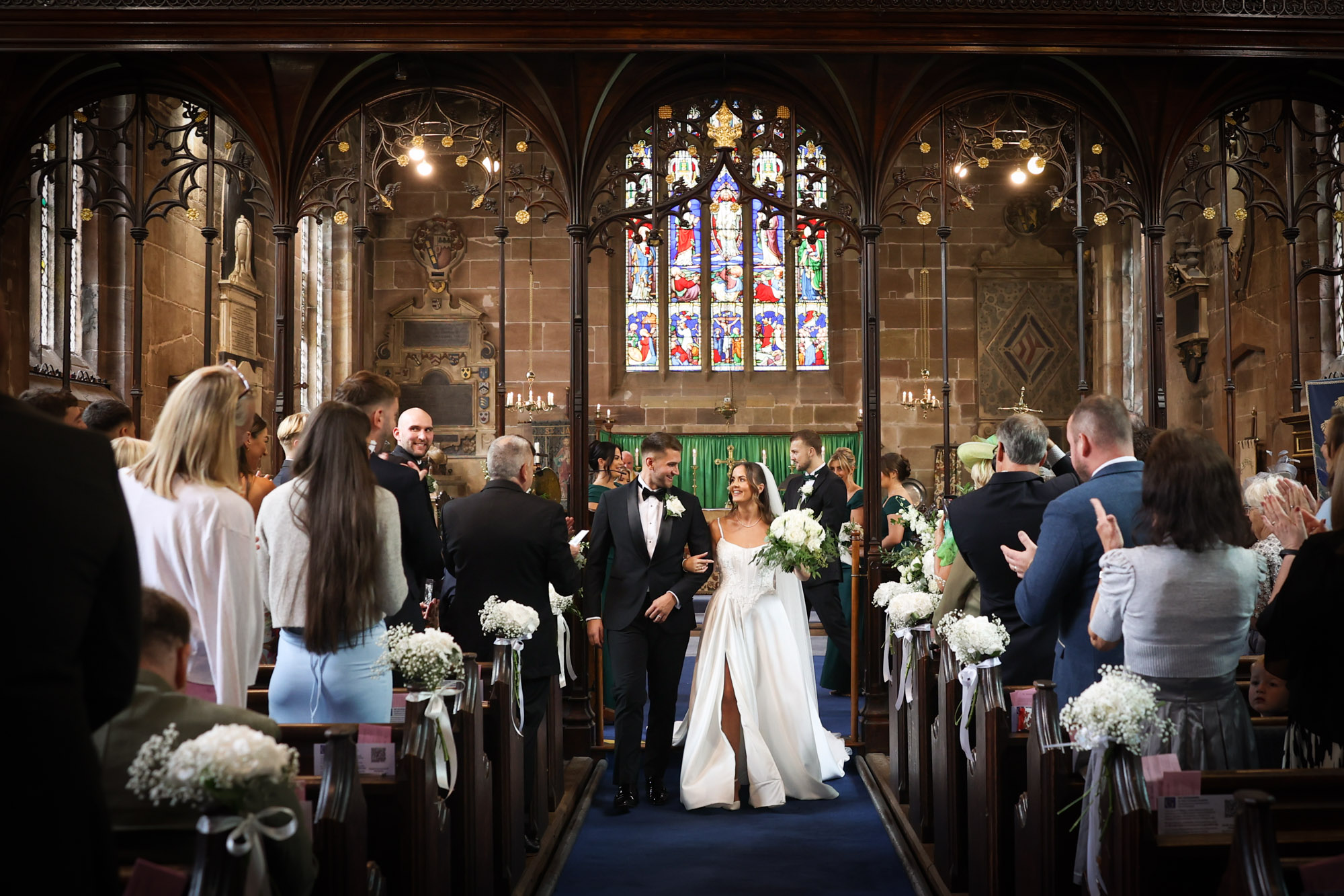 Newlywed couple walks down aisle smiling as guests applaud in historic church.