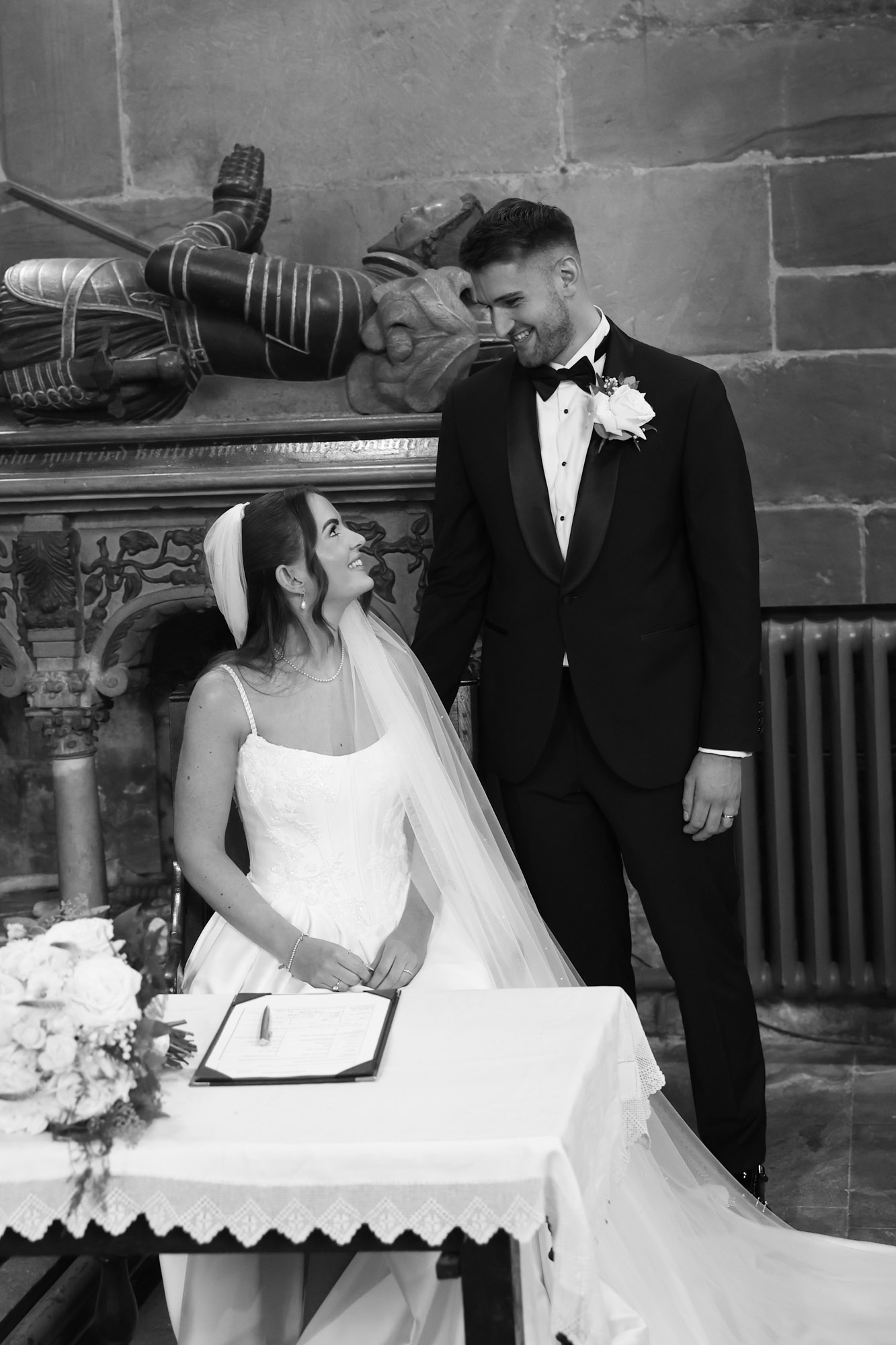 Black and white photo of seated bride signing wedding register as groom stands beside her.