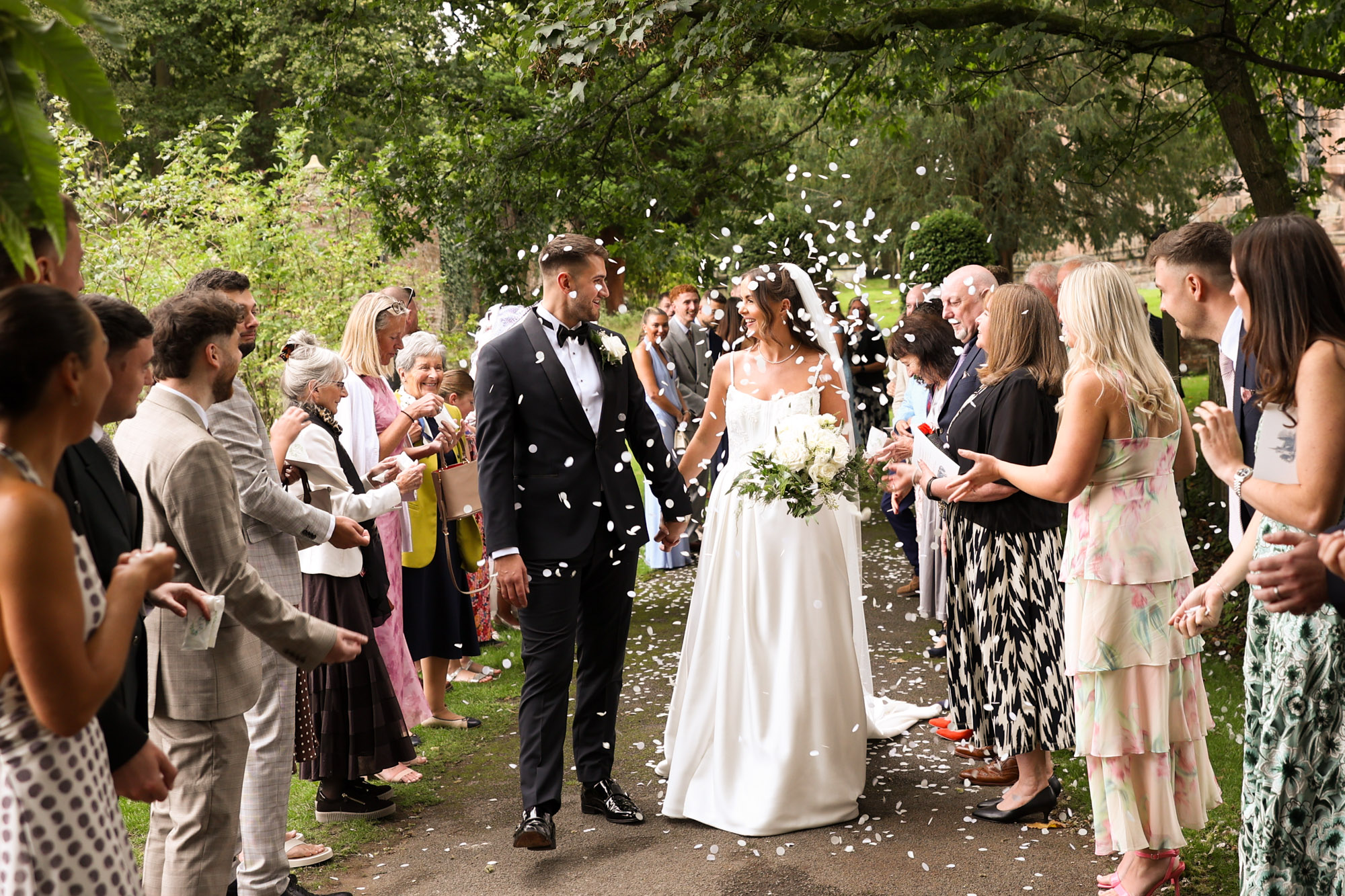 Newlyweds walk outdoors while guests throw white confetti.