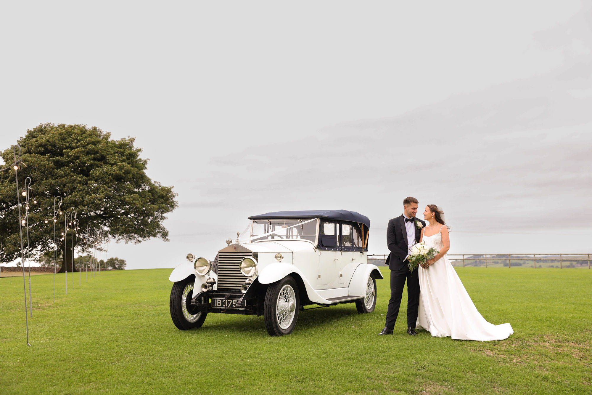 Bride and groom pose next to a white vintage car in a grassy field at Heaton House Farm.