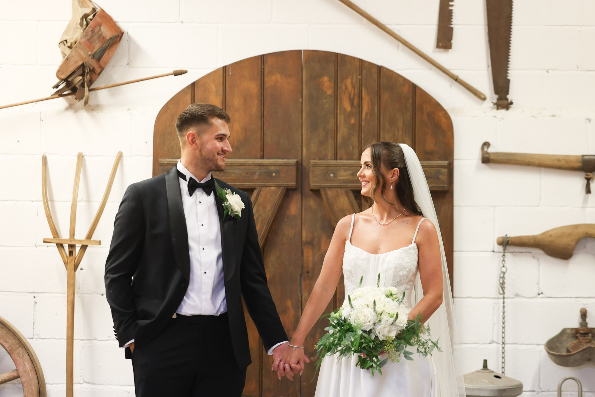 Bride and groom hold hands indoors in front of wooden barn door and farm tool decor.