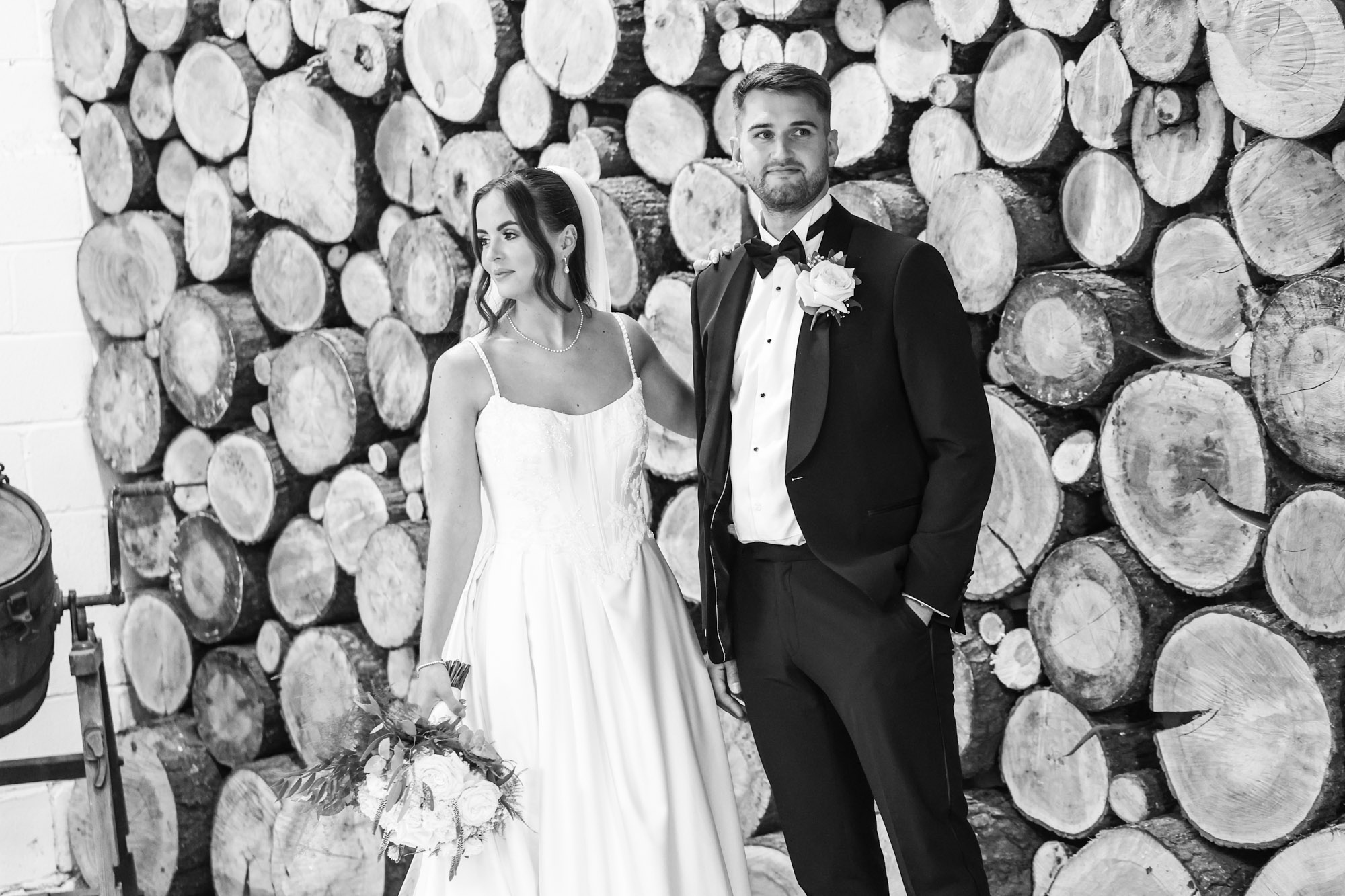 Black and white portrait of a bride and groom standing before a wall of stacked logs.