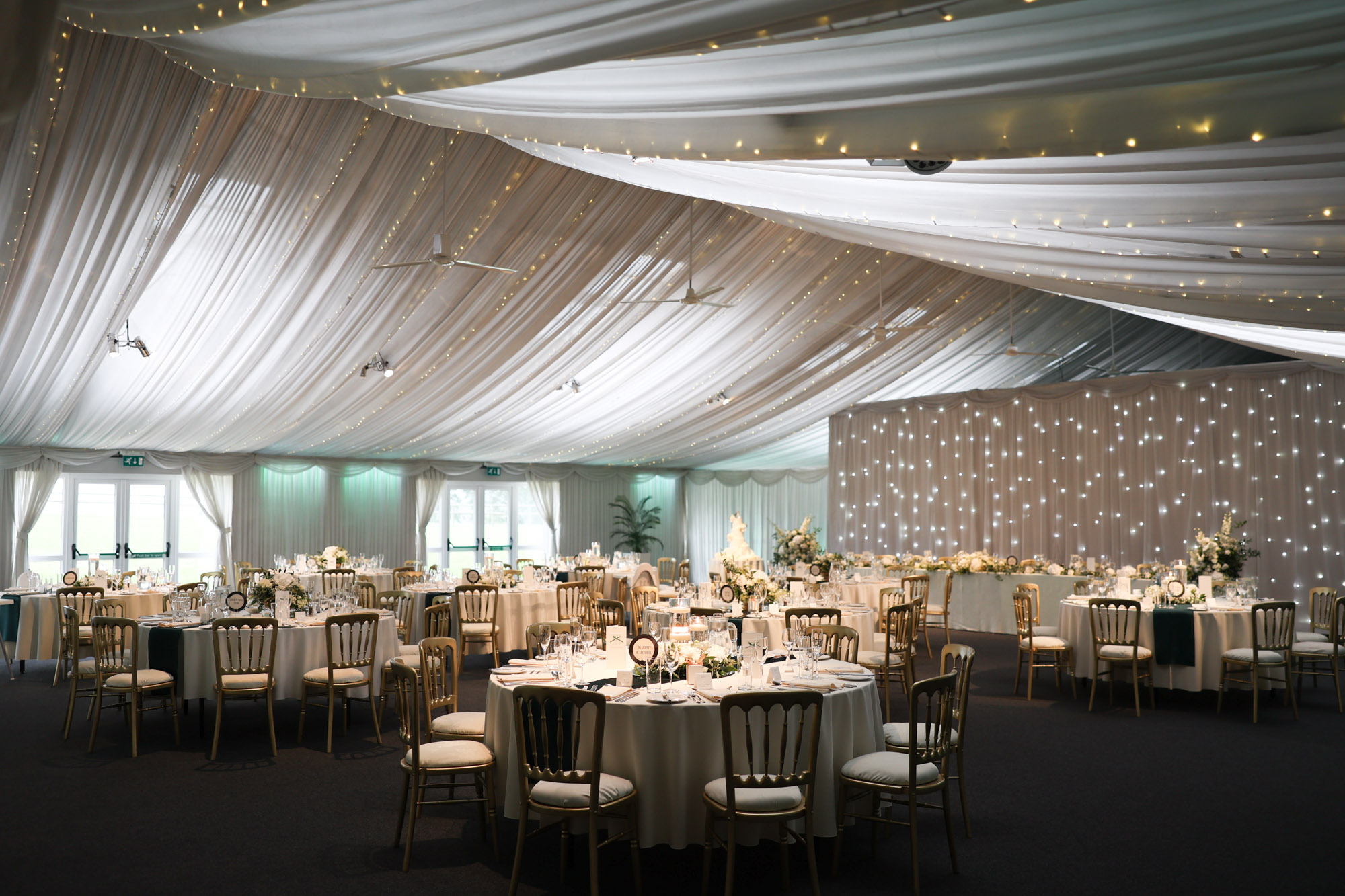 Wide interior view of a wedding reception hall with draped ceilings, fairy lights, and round dining tables.