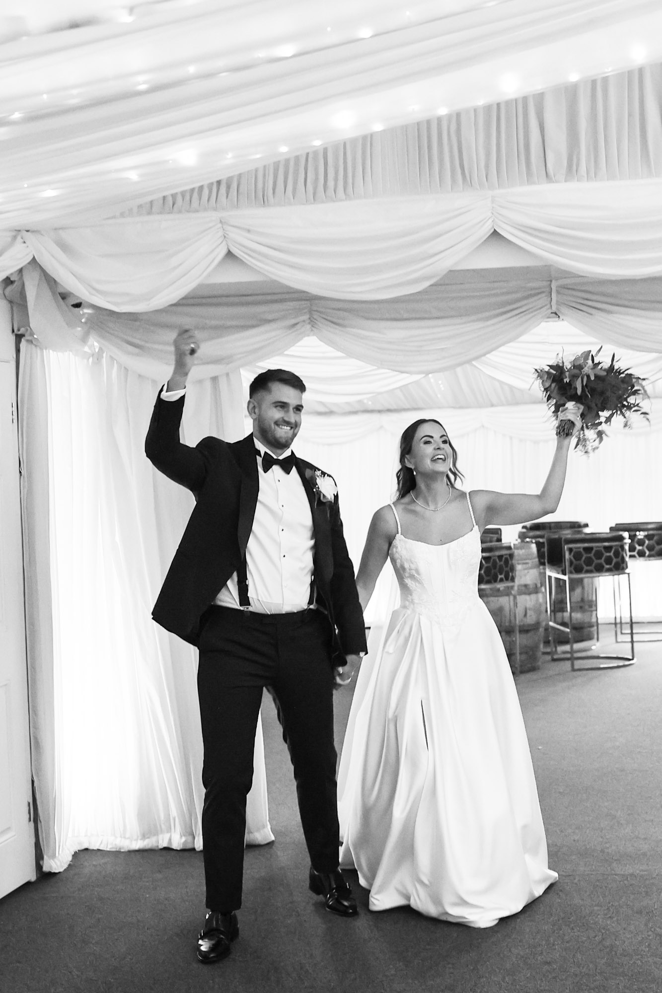 Black and white photo of a newly married couple walking up the aisle with raised arms in a white marquee.