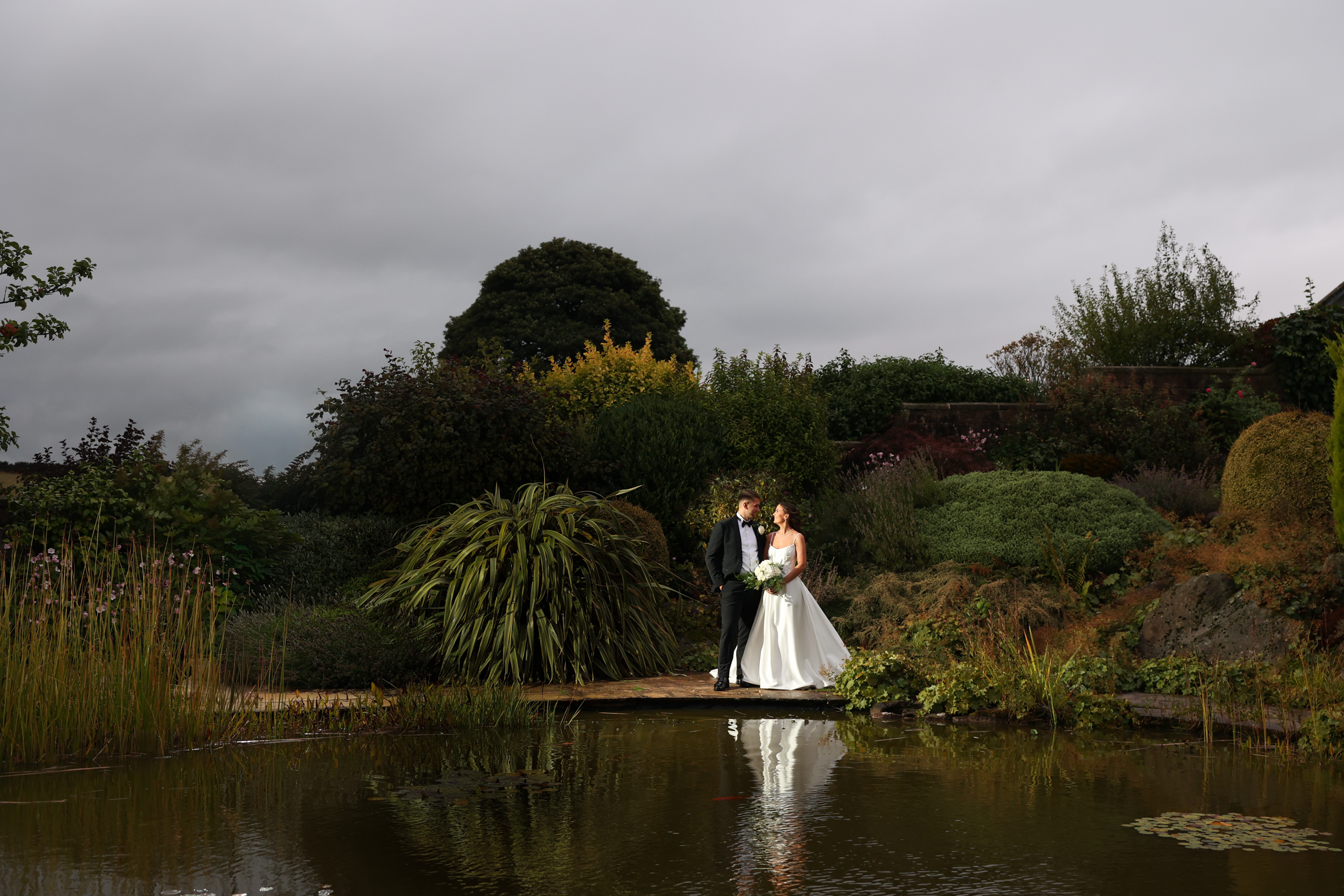 Bride and groom posing in front of a garden pond and dense shrubbery.