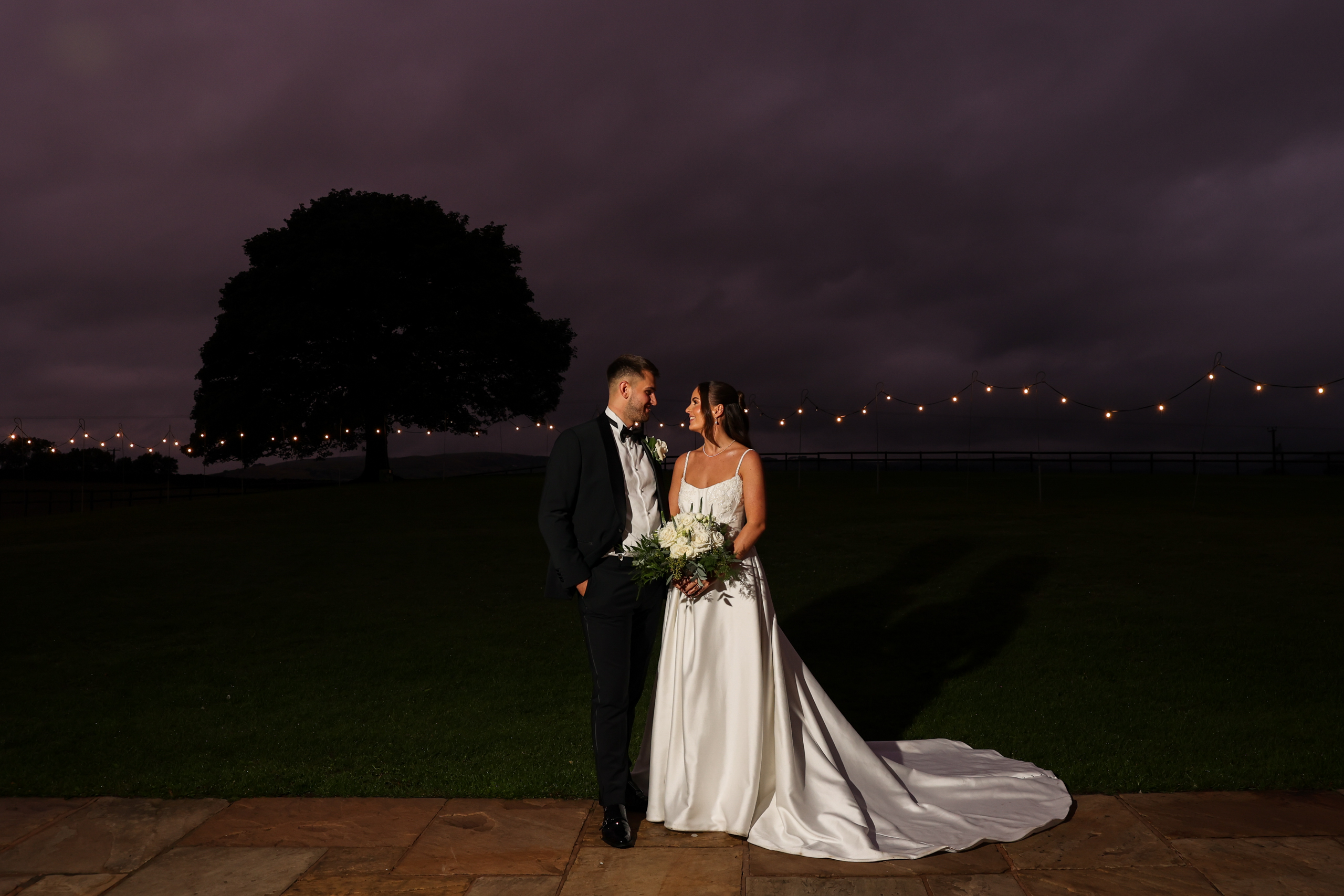 Night portrait of a bride and groom on a patio under a dramatic purple and dark cloudy sky, with string lights.