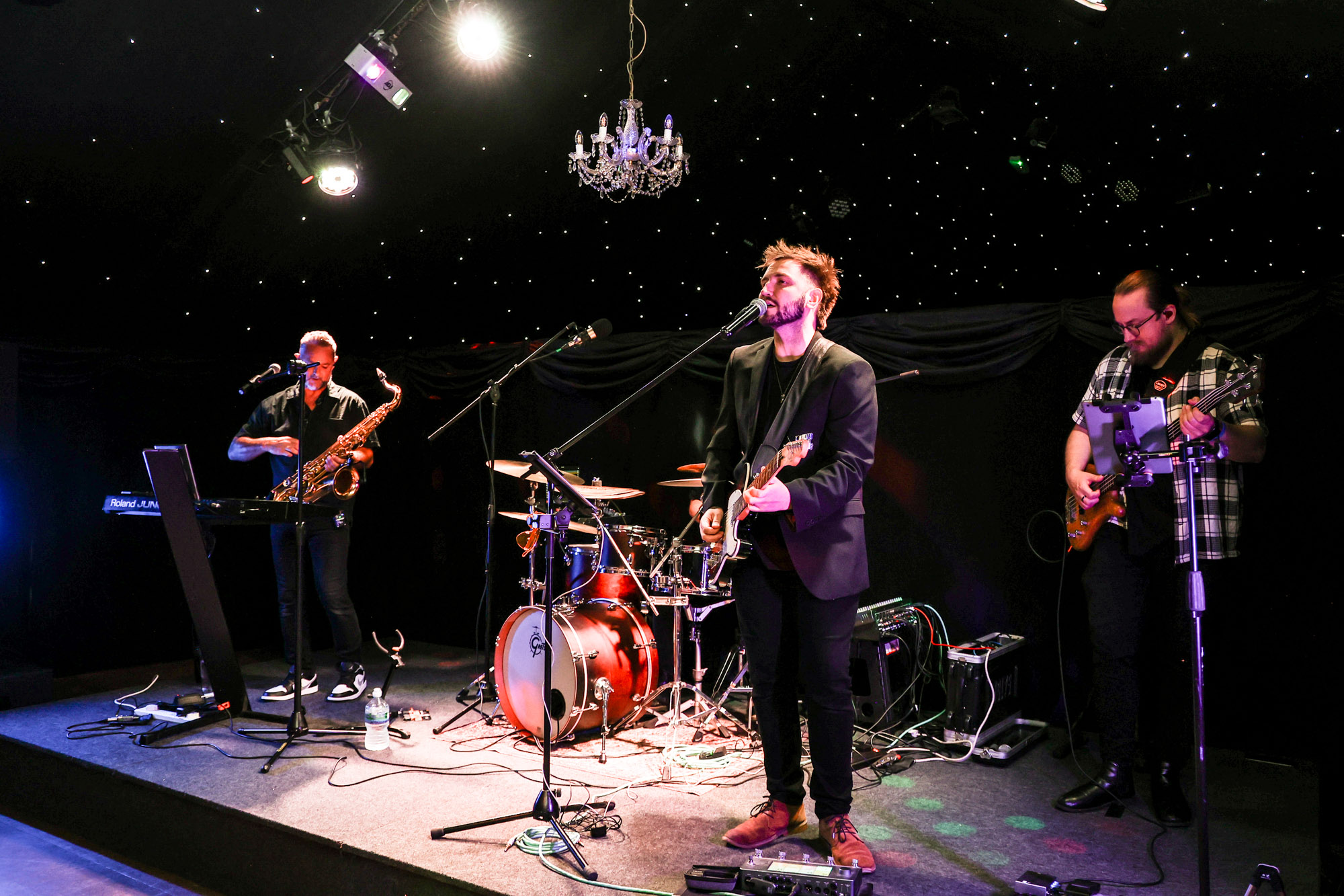 Wide shot of a four-piece band (vocalist, saxophonist, drummer, bassist) performing live on a stage in a white marquee.