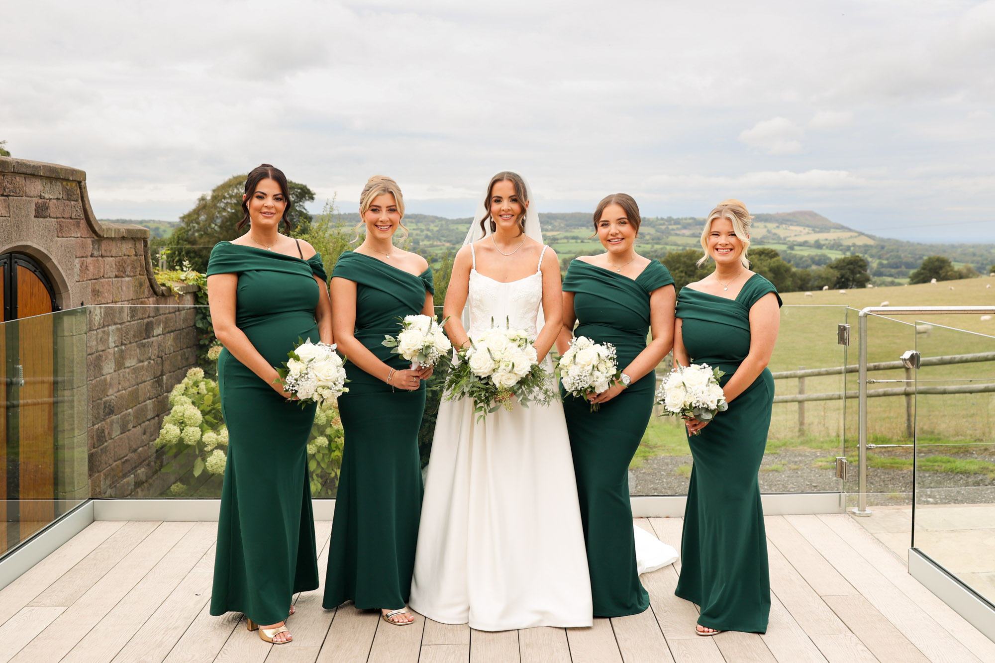 Bride in white gown poses outdoors with four bridesmaids in dark green dresses.