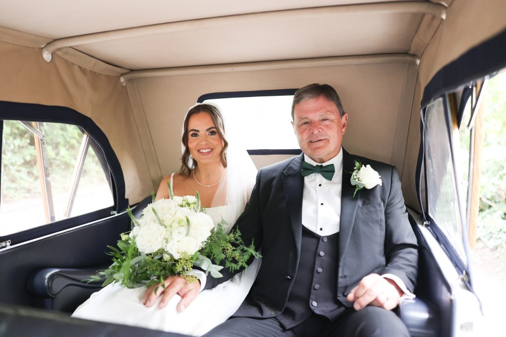 A bride with a veil and white rose bouquet sits with her father in a tuxedo inside a vintage wedding car.
